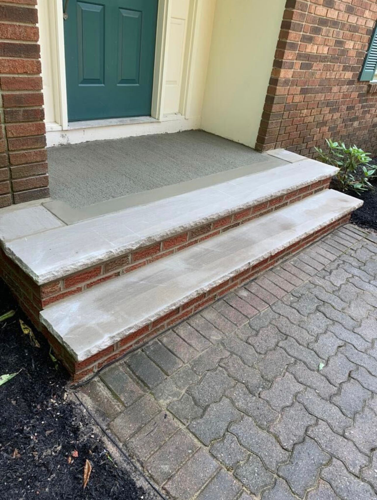 Brick steps leading up to a green door, with gray stone treads and a brick facade, on a brick paved walkway.