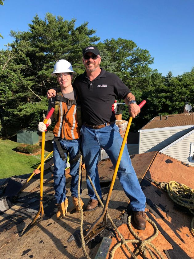 Two people stand on a roof, one in safety gear. They are holding tools; sunny day, trees in background.