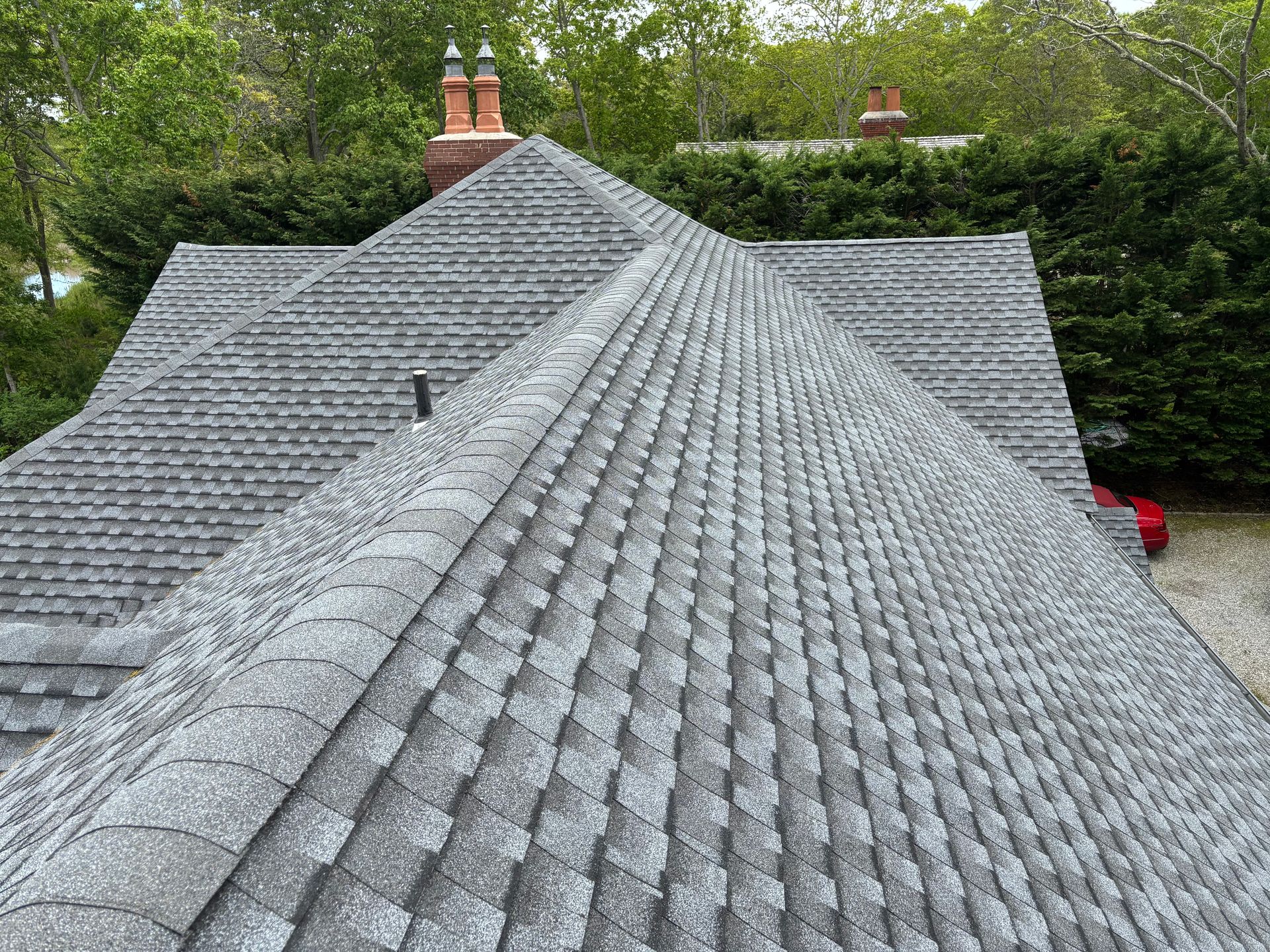 Gray shingled roof of a house, set among green trees. Two chimneys visible.
