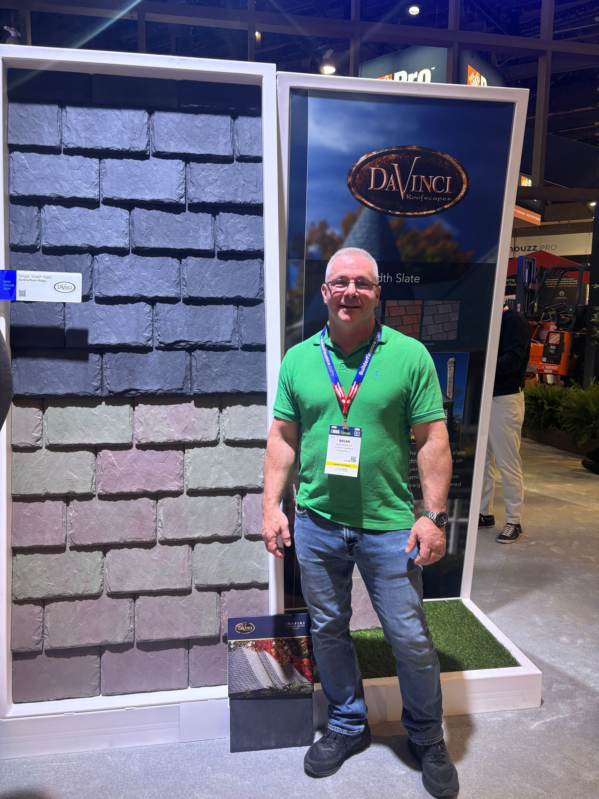 Man in green shirt stands by a display of roofing tiles at a trade show.