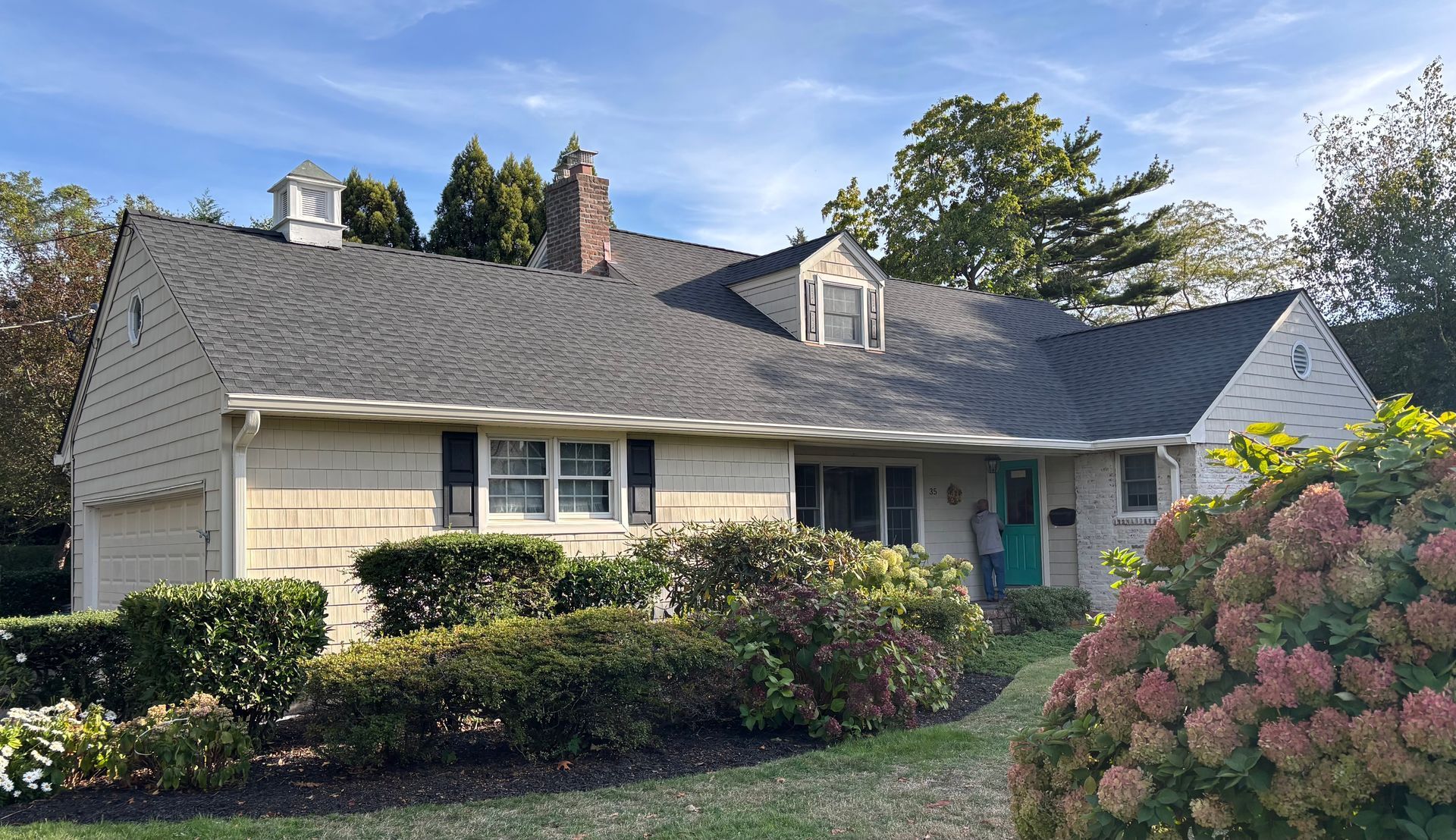 Beige house with a dark roof and a turquoise door, surrounded by bushes and trees under a blue sky.