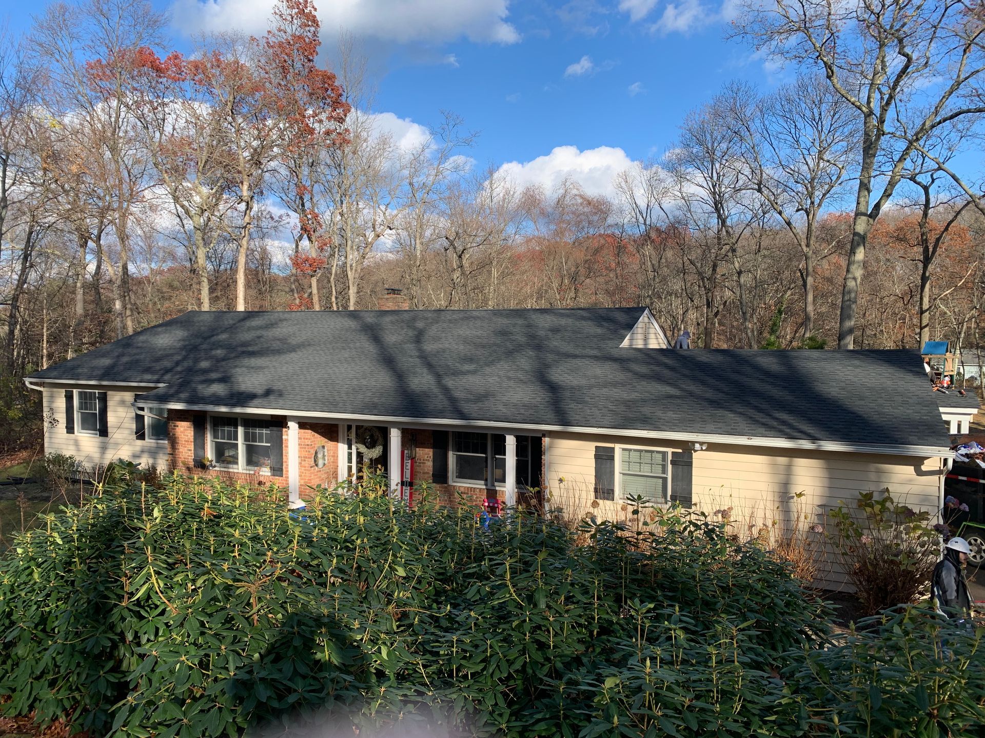 Ranch house with black shingle roof, beige brick exterior, and lush green bushes in front. Fall trees in the background.