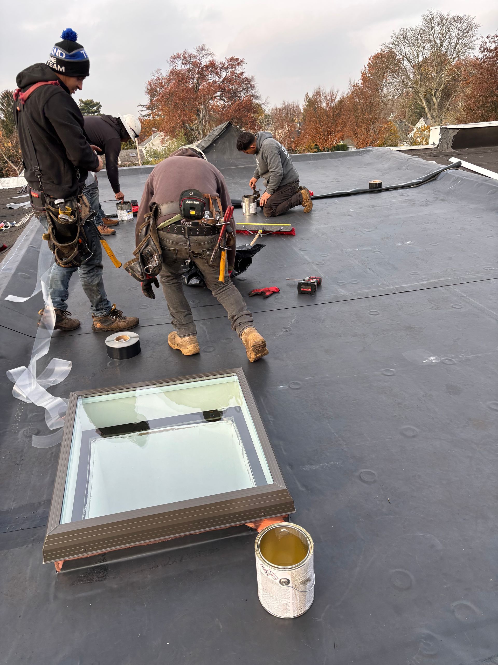 Roofers working on a flat roof, installing a skylight. Tools and supplies are visible, outdoors.
