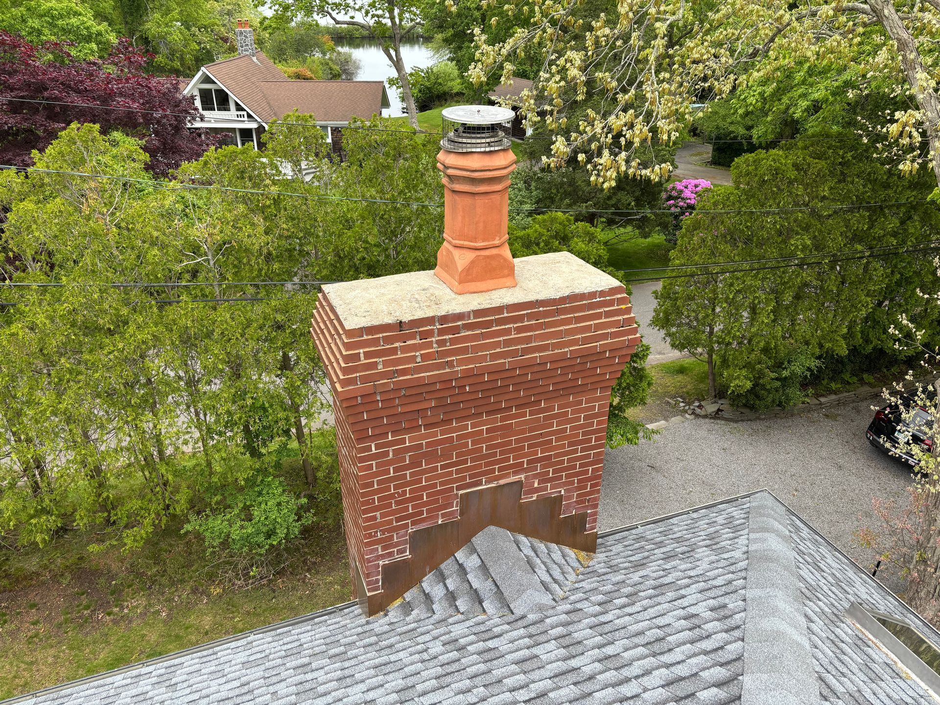 Brick chimney on a gray shingled roof, topped with a terracotta flue and a metal cap, surrounded by greenery.