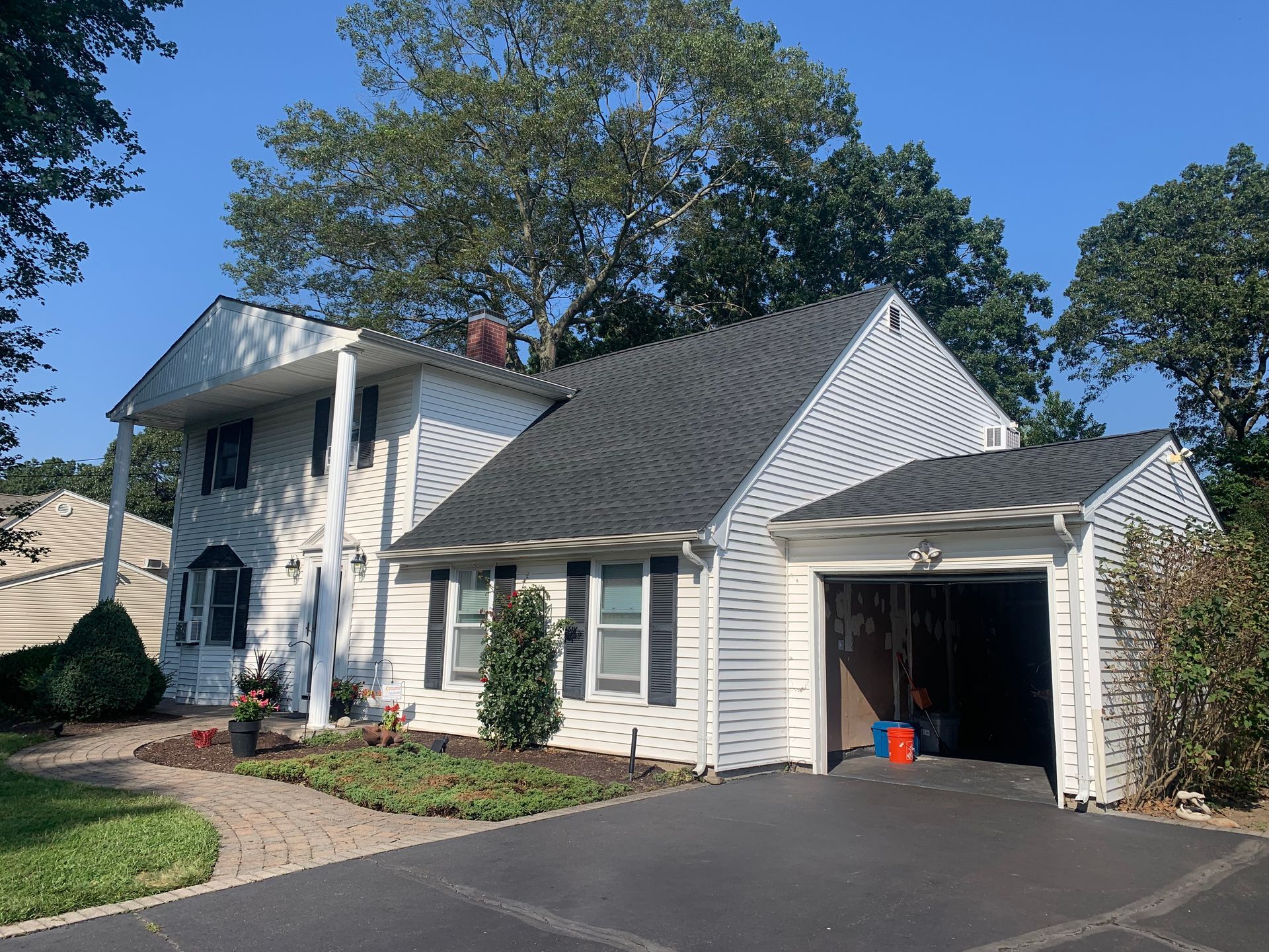 White two-story house with black roof, attached garage, and paved driveway. Surrounded by trees and greenery.