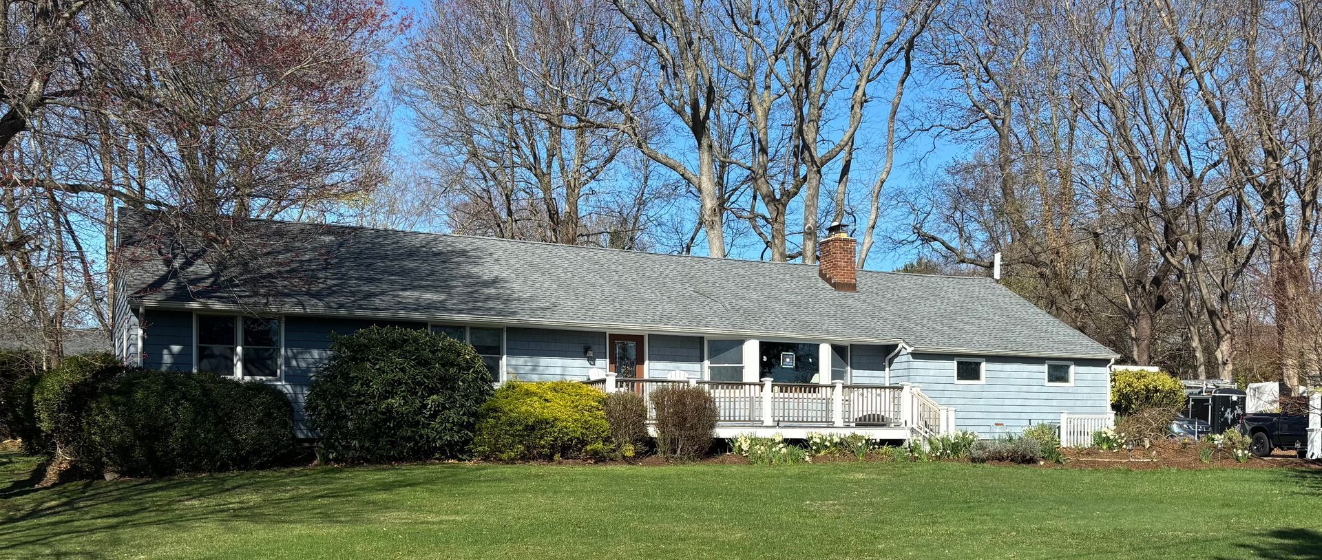 A light blue ranch house with a gray roof and a brick chimney surrounded by trees on a sunny day.