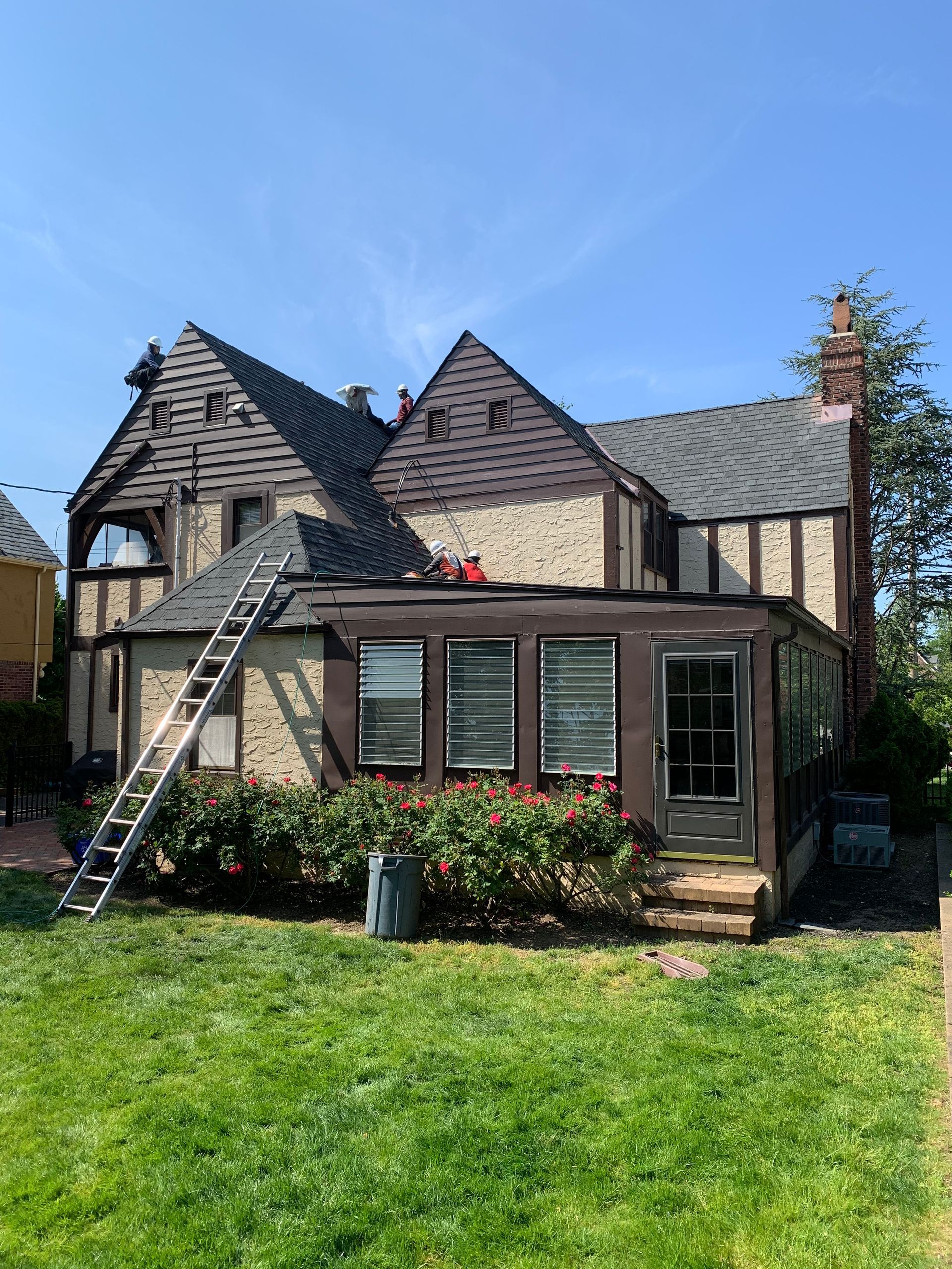 House with brown trim, roofers on roof, ladder, and lush green lawn.