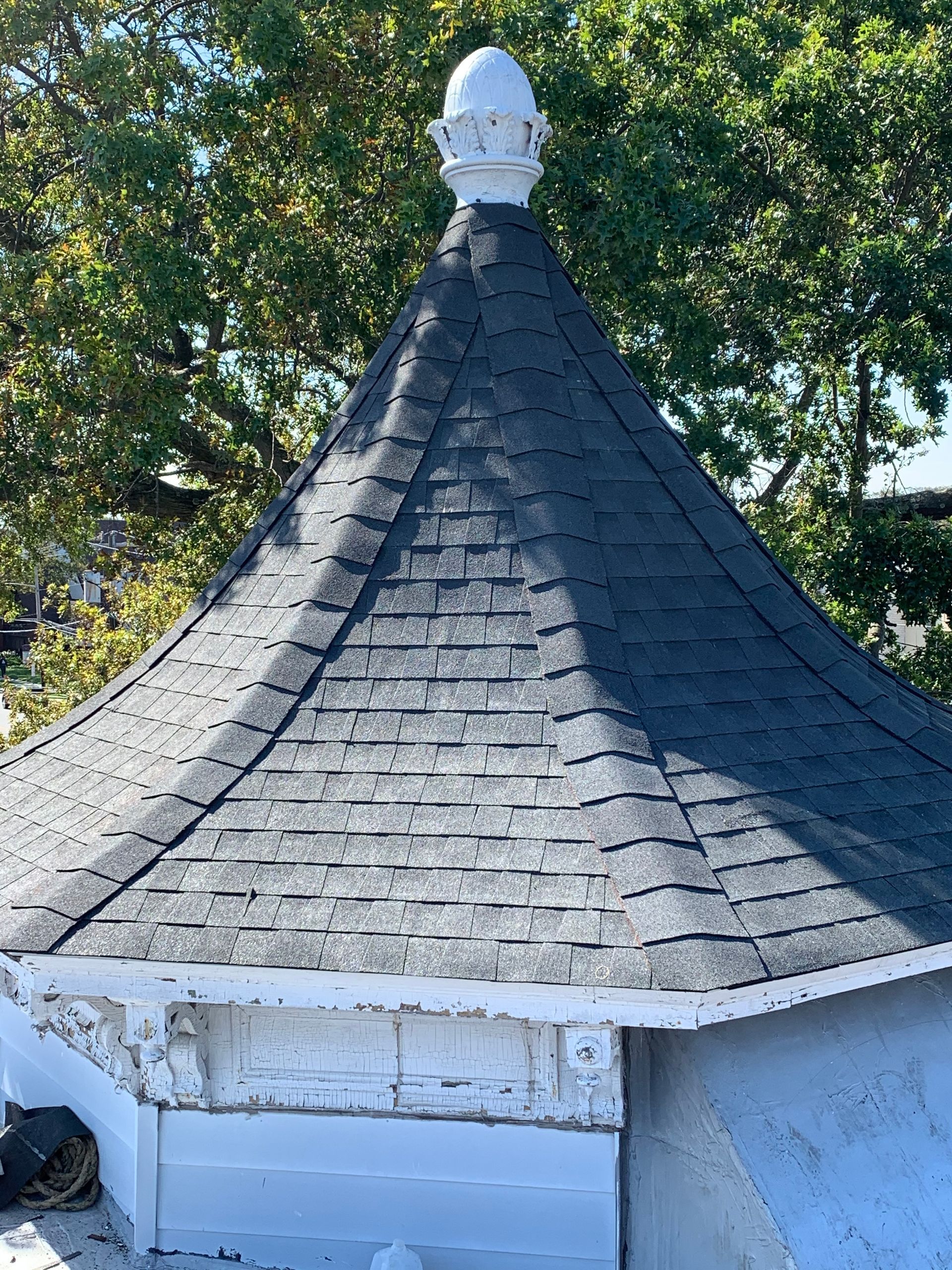 A dark shingled, cone-shaped roof with a white decorative top, set against a background of green trees.