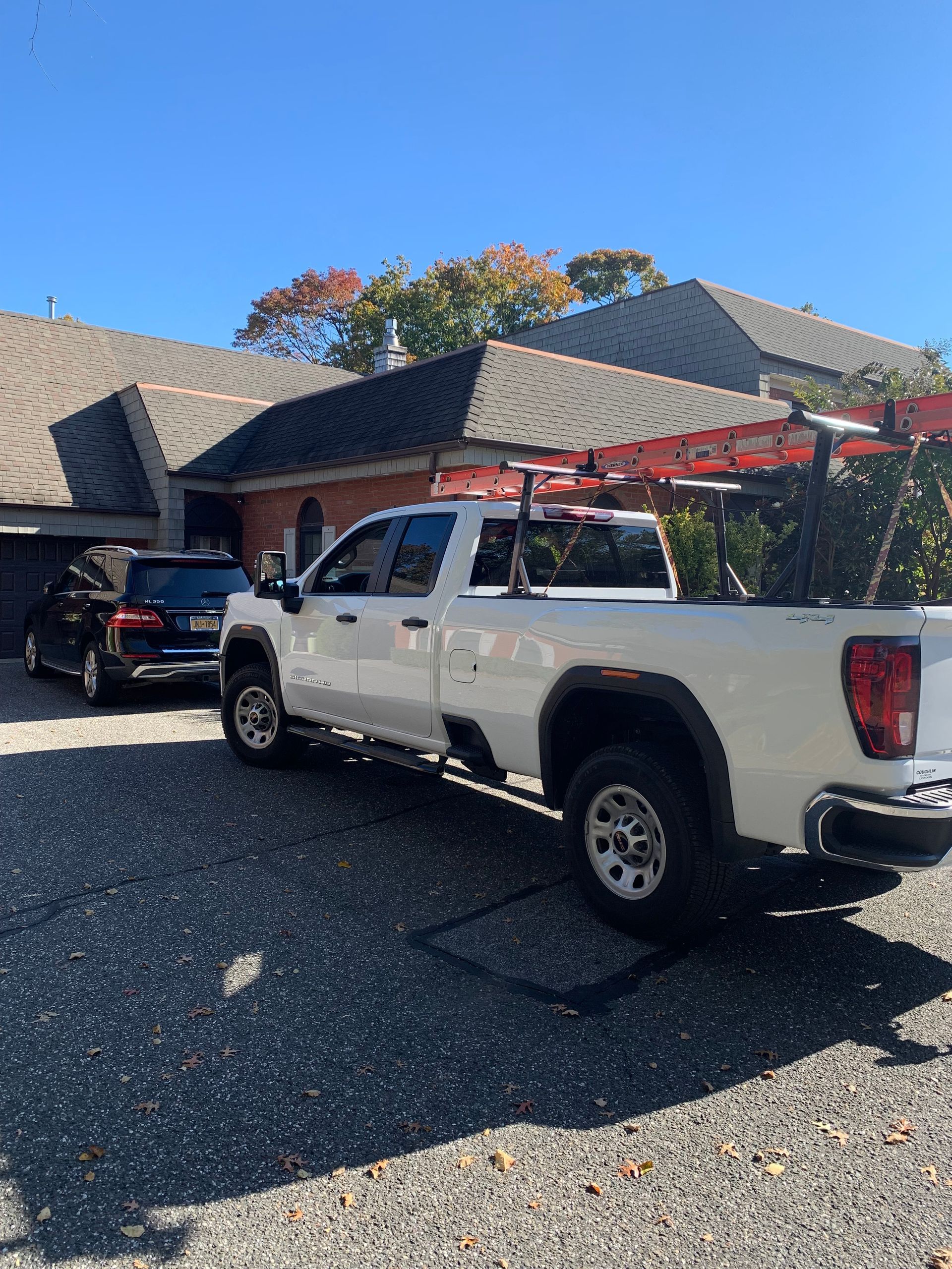 White pickup truck parked in front of a house, with ladder on top. A black SUV is parked beside it.