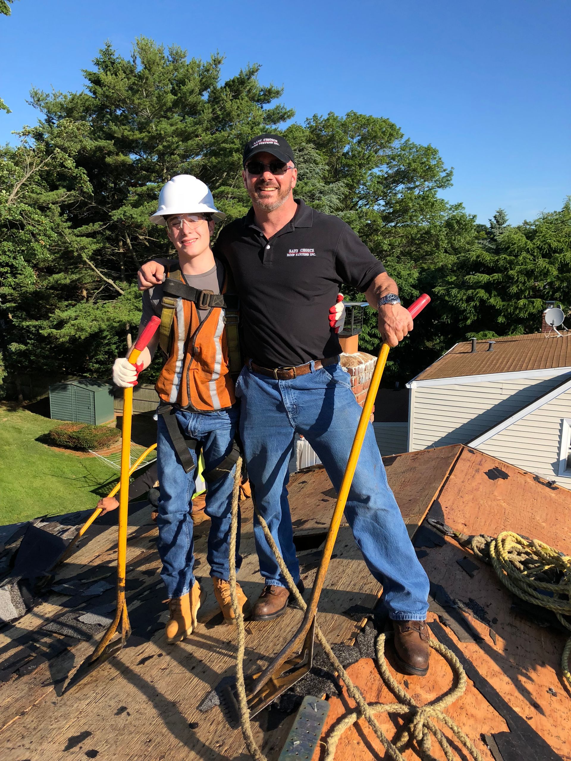 Two men on a rooftop: one in a harness, hardhat, holding a rake, and the other in a black shirt, holding a rake. Blue sky.