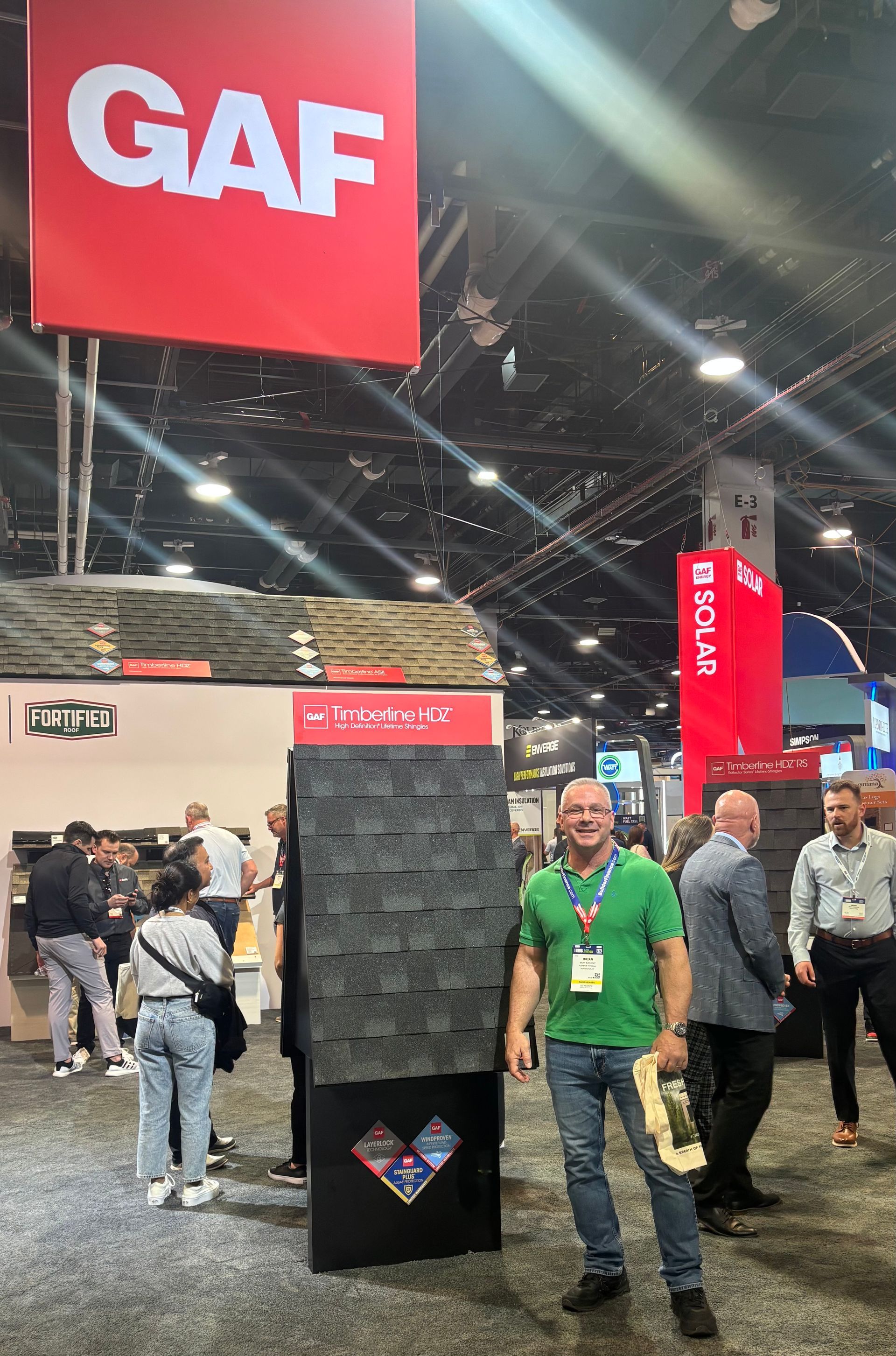Man in green shirt stands by GAF roofing display at a trade show, other people visible.