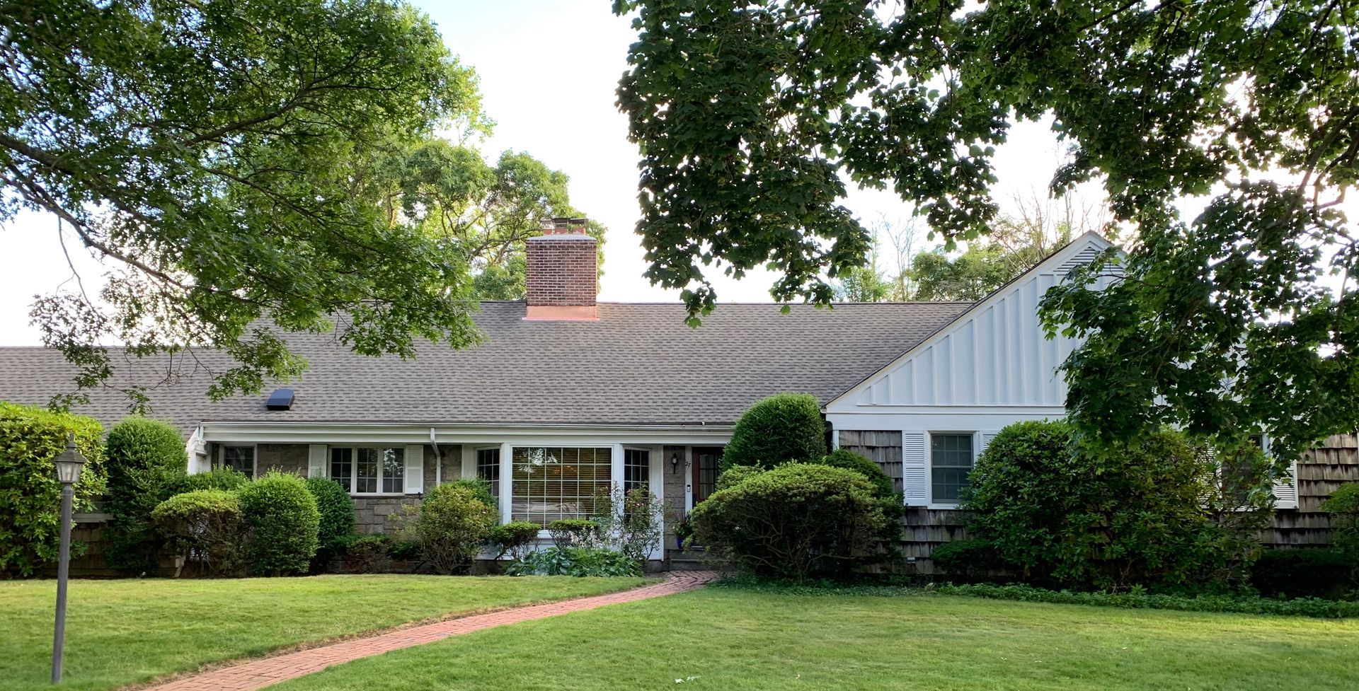 Ranch-style house with gray roof, white accents, and green lawn, framed by leafy trees.
