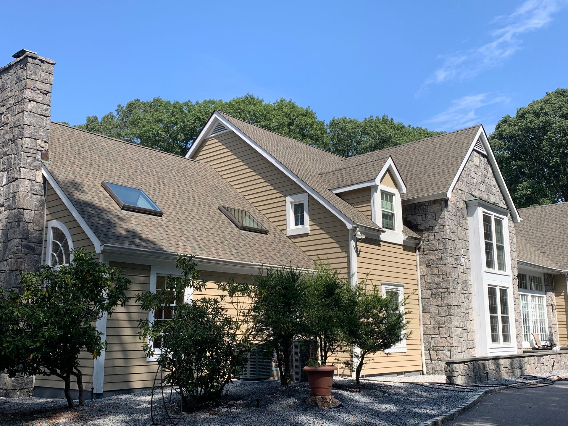 House with stone chimney and accents, beige siding, and a tan roof against a blue sky.