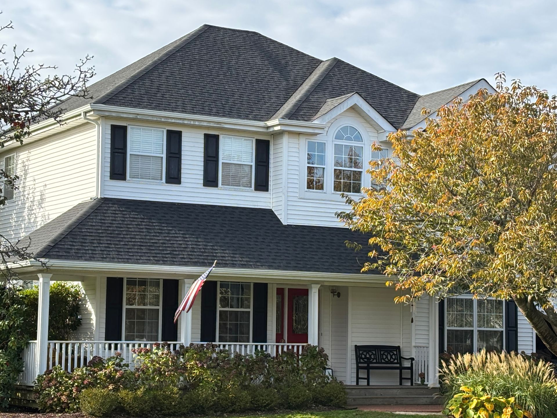 Two-story white house with black shutters and roof; front porch, red door, and American flag.