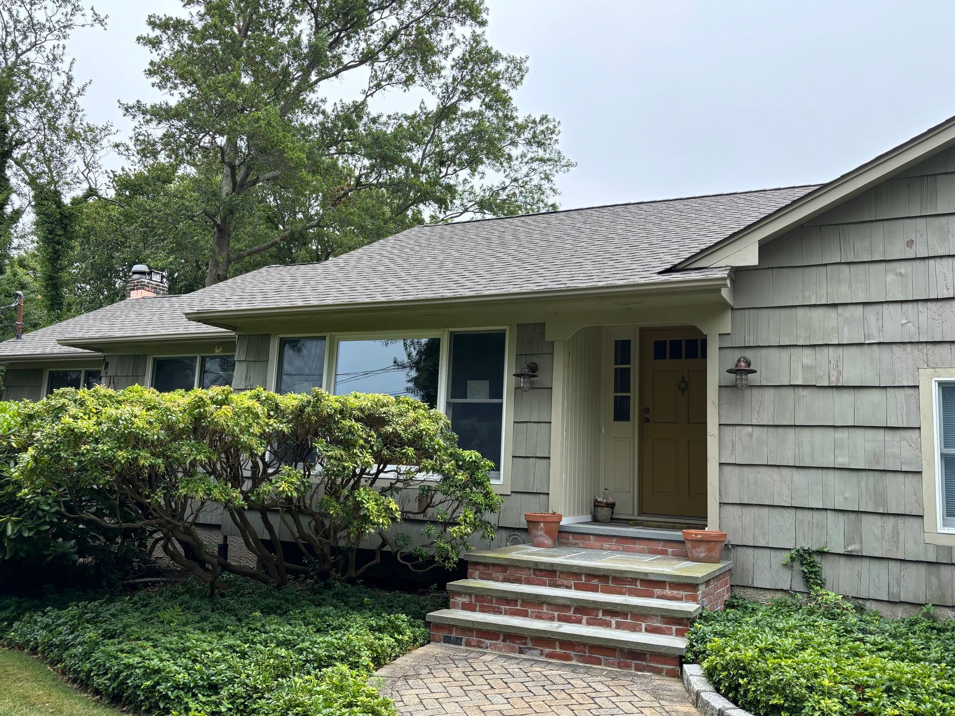 A light gray house with a yellow door, red brick steps, and a lush green bush in front.