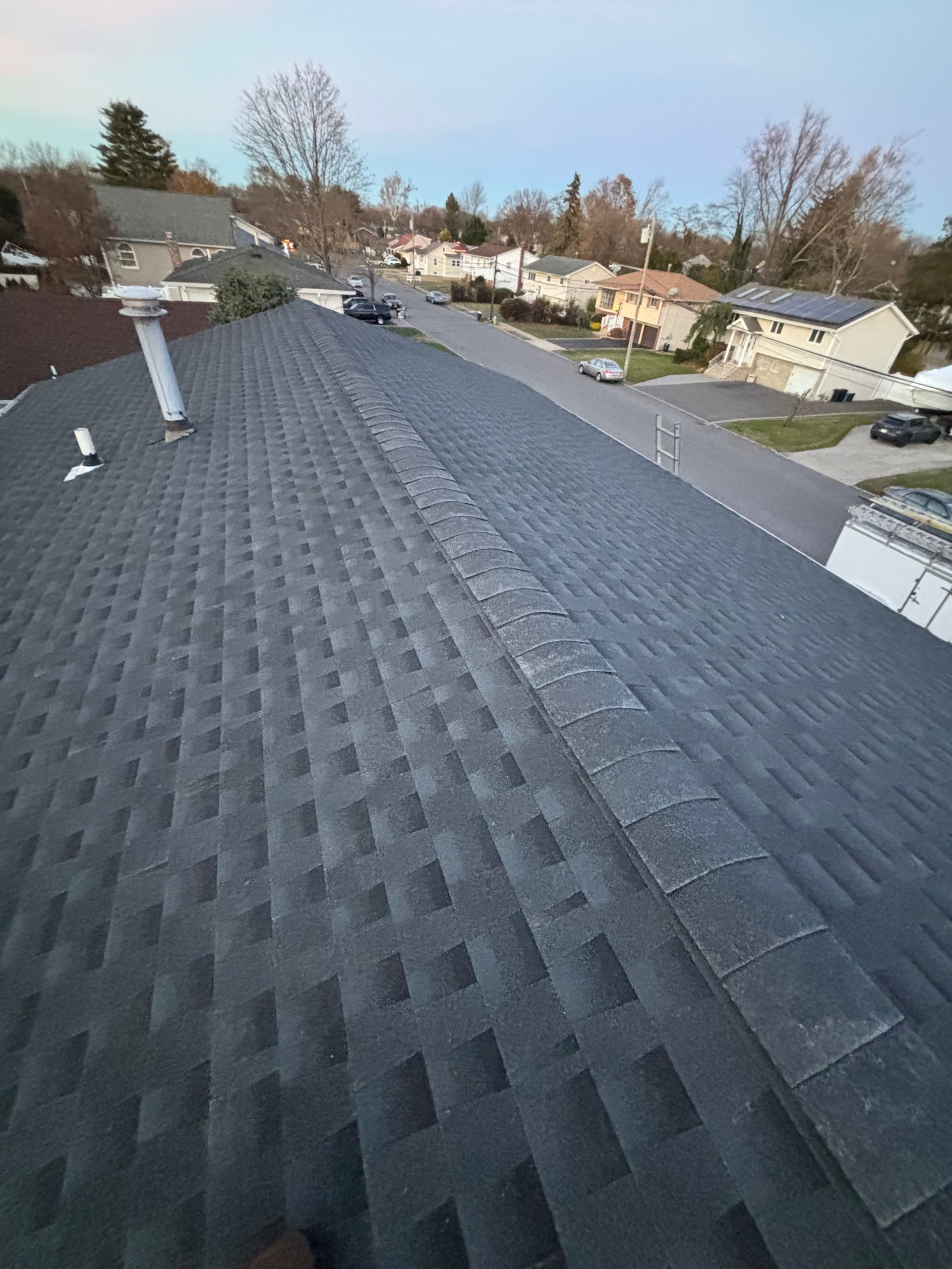 View of a dark asphalt shingle roof. A street and houses are in the background under a blue sky.