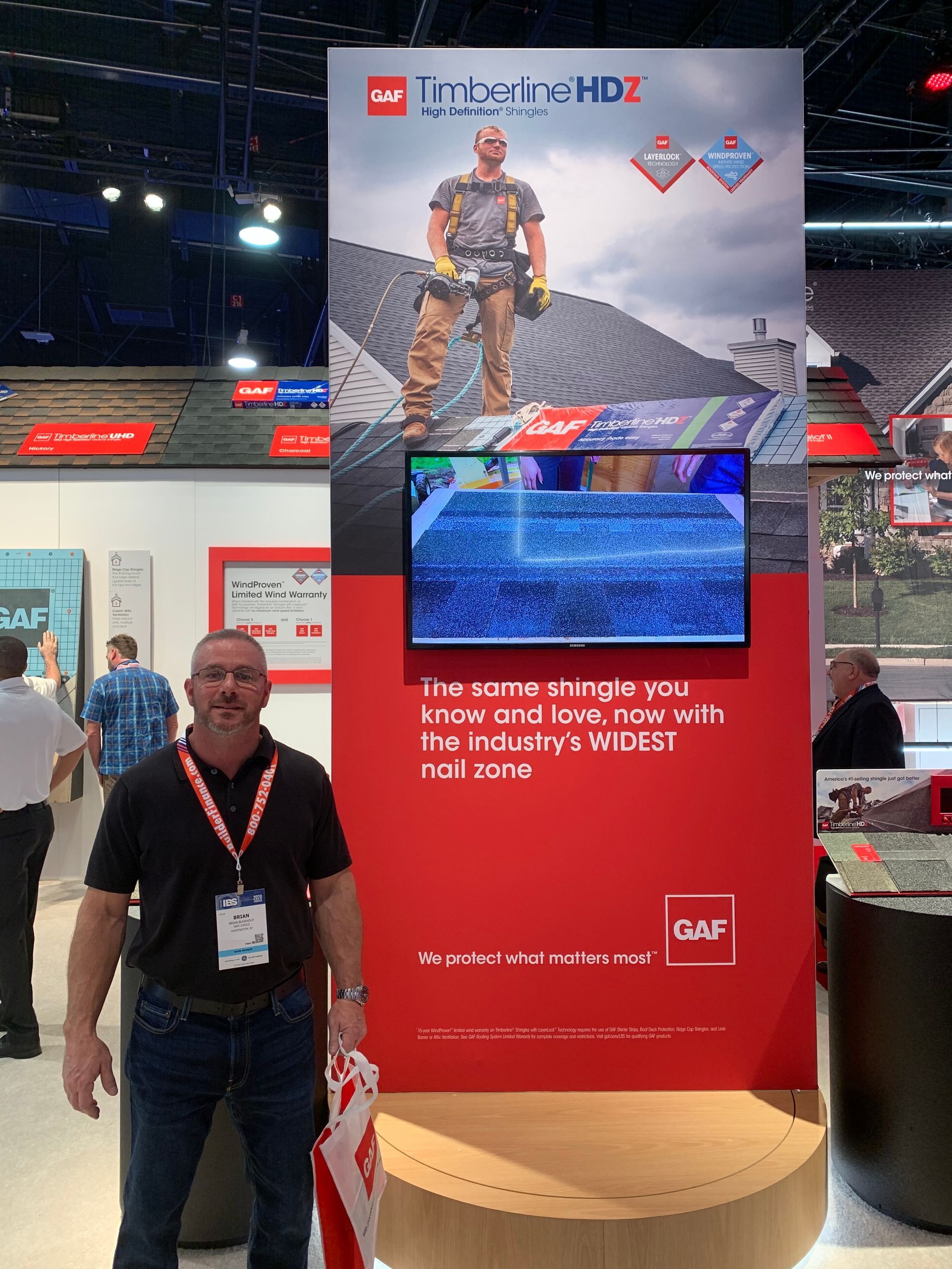 Man stands near a GAF Timberline HDZ display at a trade show. The display shows a man on a roof, with shingles.