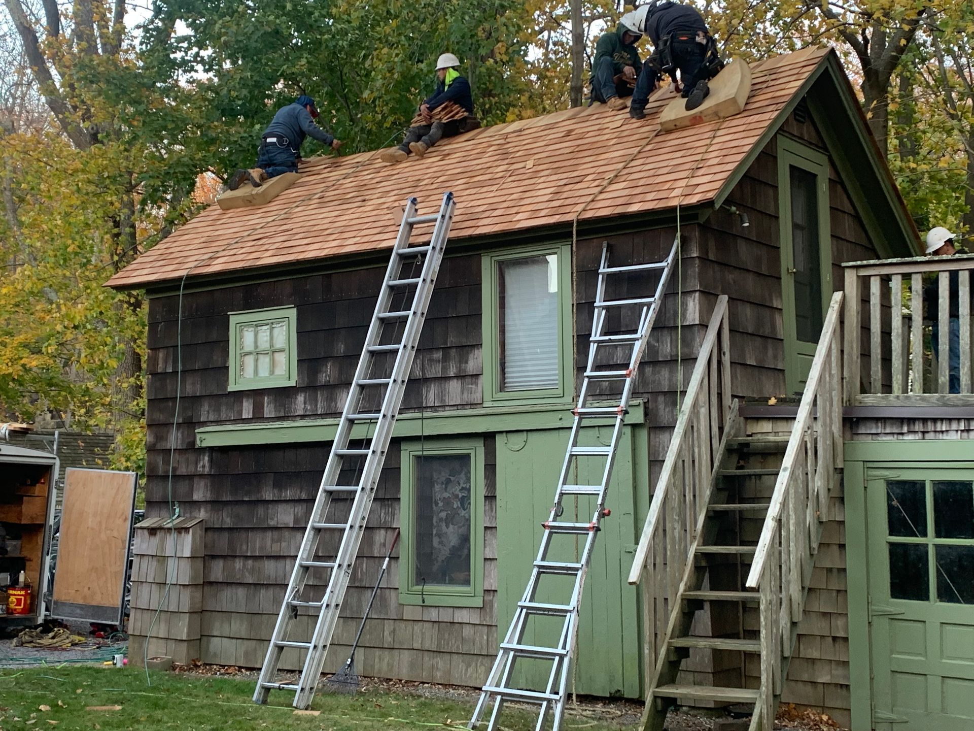 Workers re-roofing a small, weathered brown building with green trim; two ladders lead to the roof.