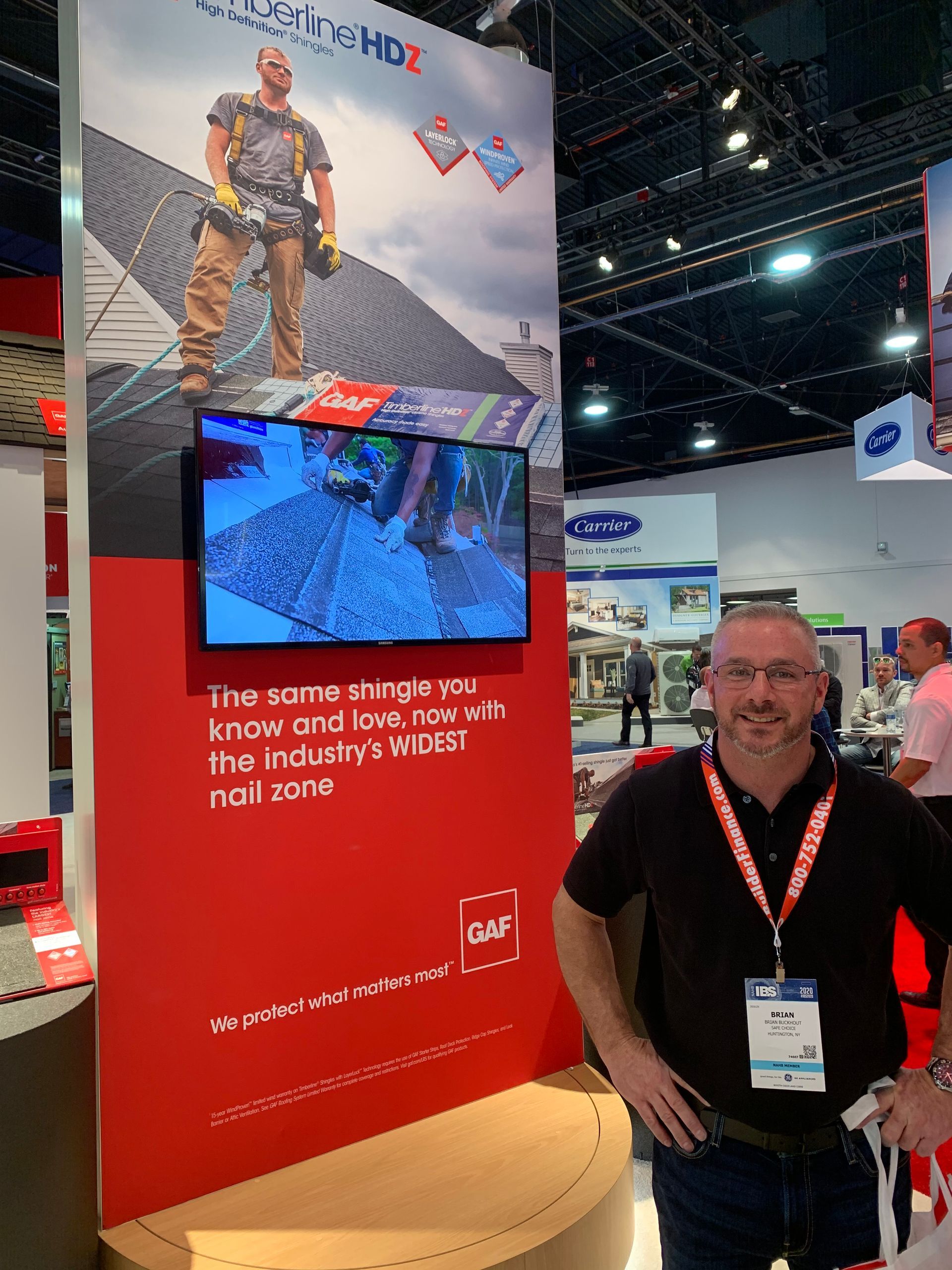 Man in front of a red booth with a roofing advertisement and a monitor displaying a worksite.