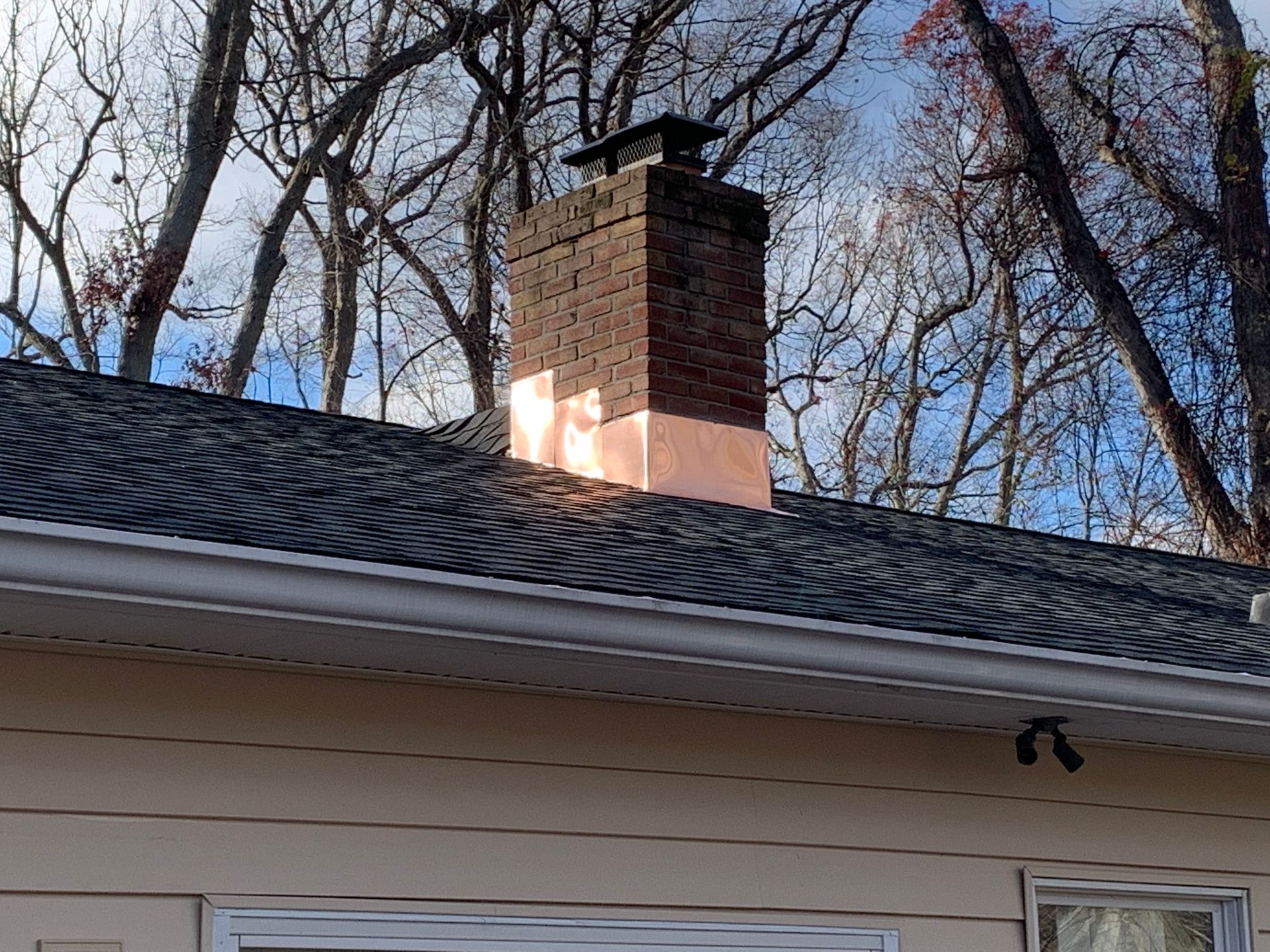 Brick chimney with copper flashing on a roof with dark shingles and light siding.