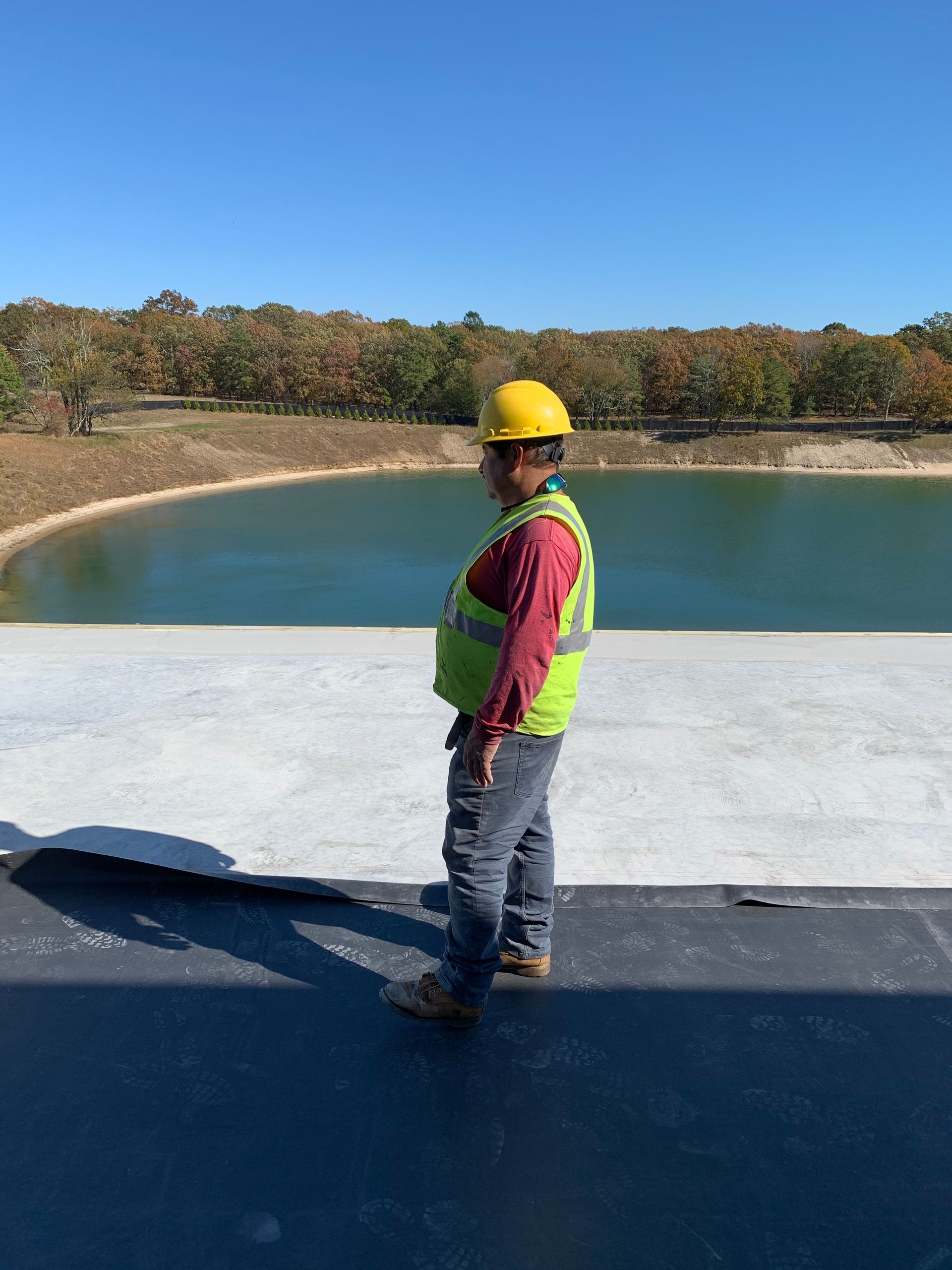 Construction worker in hard hat and vest stands on a roof, overlooking a lake.