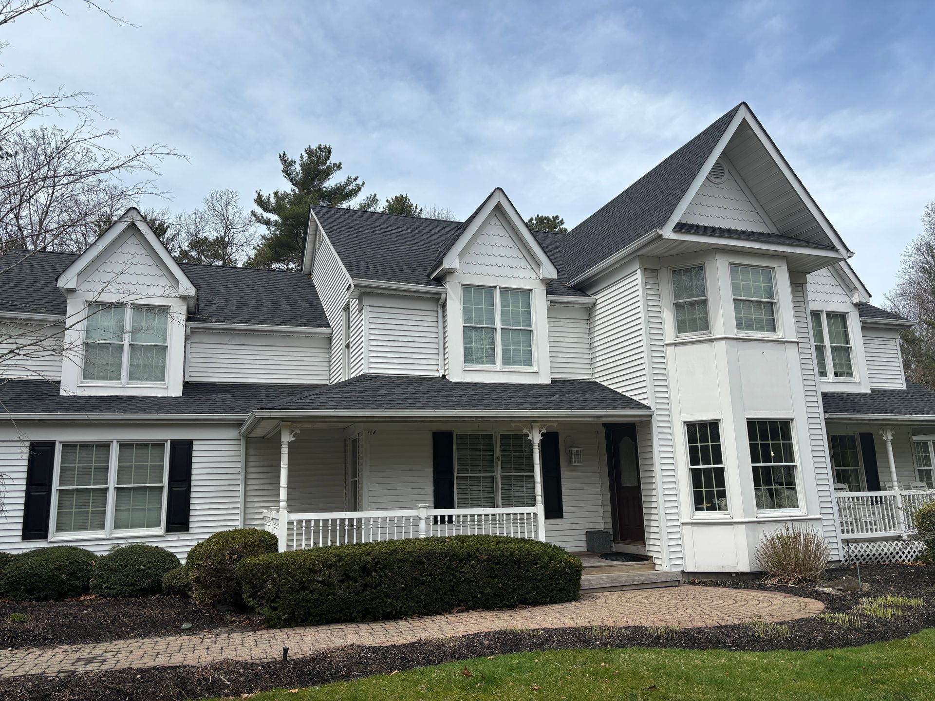 White house with black shutters, dark roof, multiple dormers, and a small porch, cloudy sky.