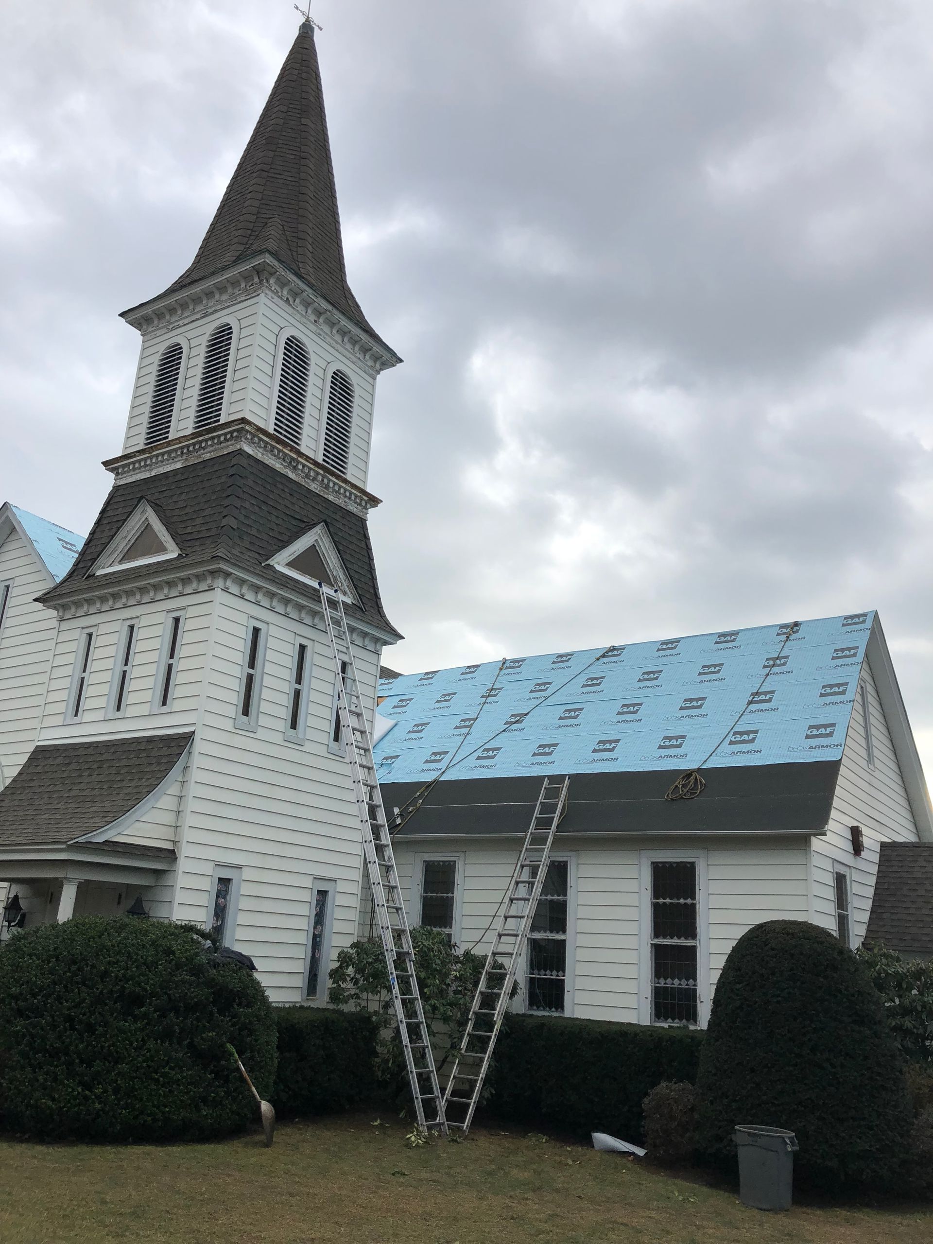 White church building with steeple, ladders against roof, blue underlayment, cloudy sky.