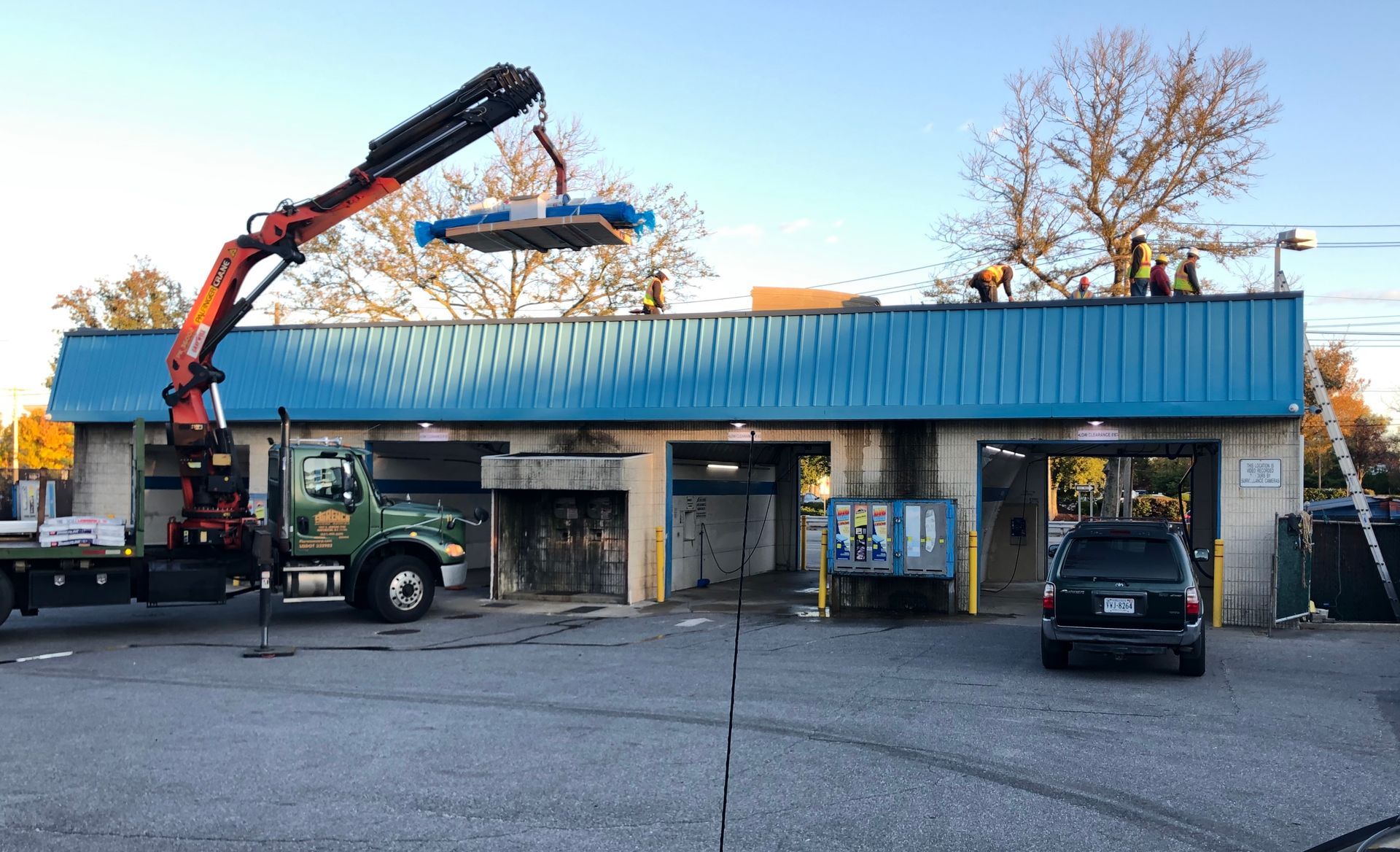 A crane lifting equipment onto a blue-roofed building where workers are present. A dark car is parked nearby.