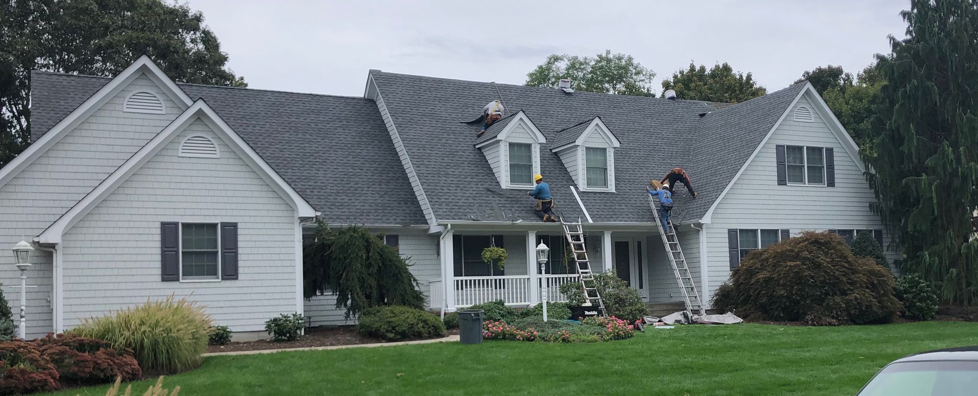 Workers on a roof, installing shingles on a white house. Two ladders propped up against the house.