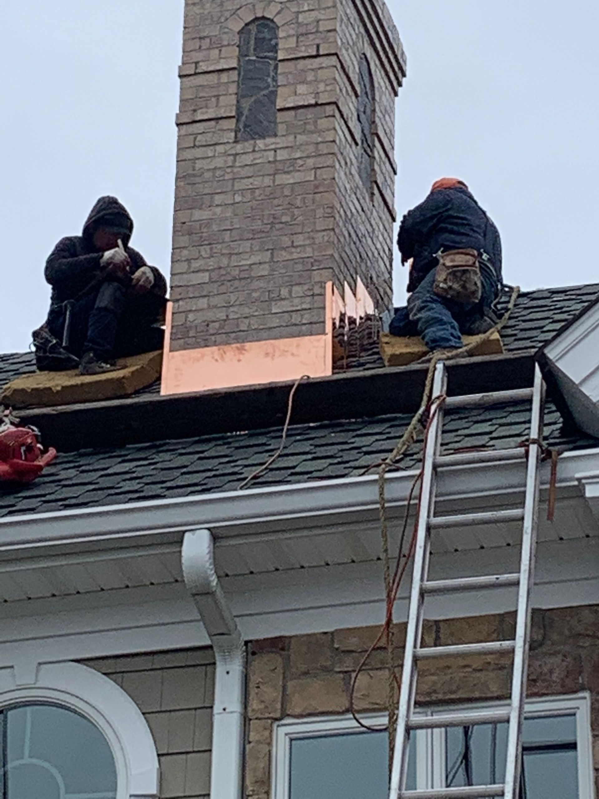 Two workers on roof near chimney installing copper flashing.