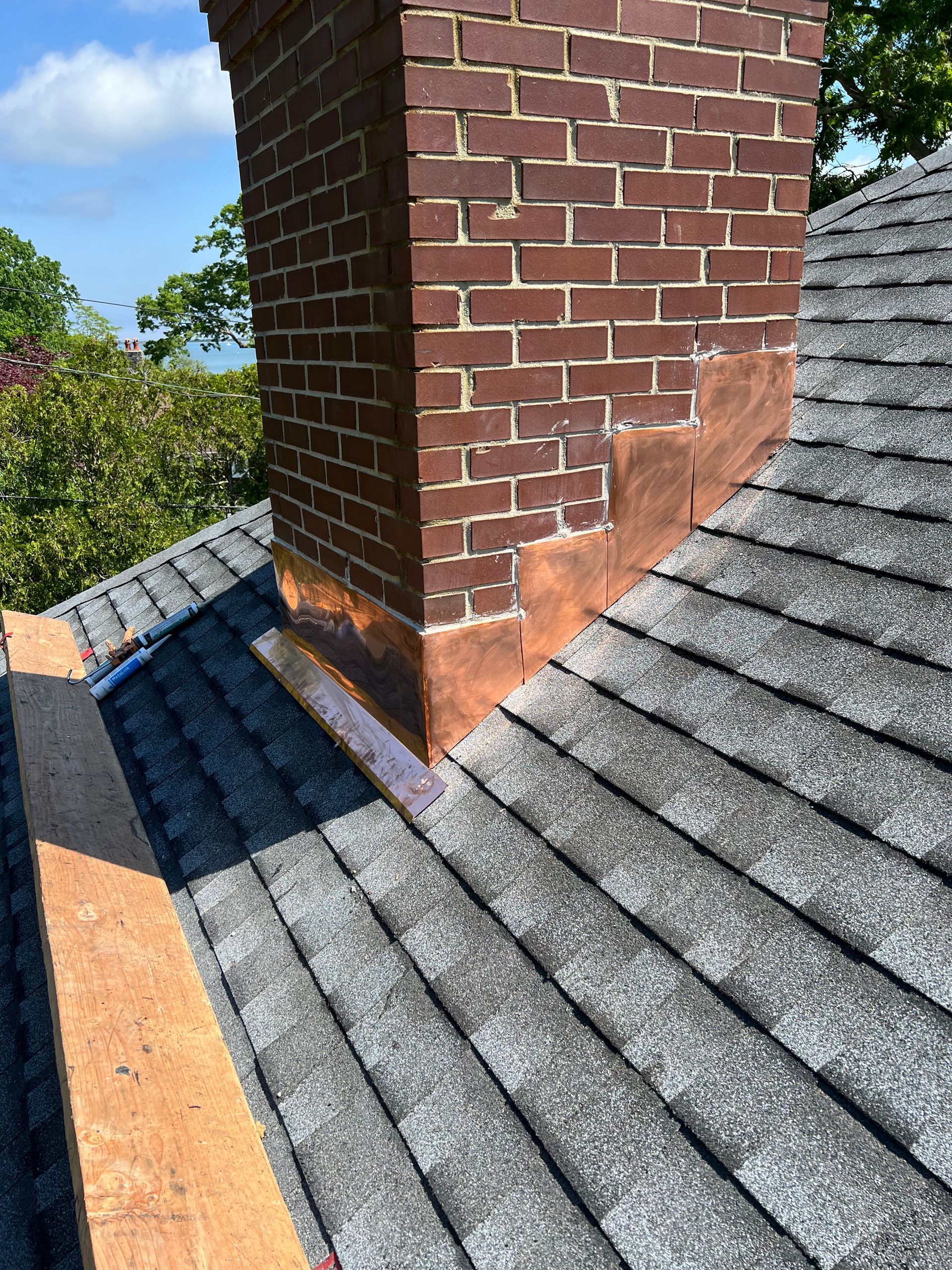 Brick chimney on a shingled roof with copper flashing visible. Cloudy sky and greenery in the background.