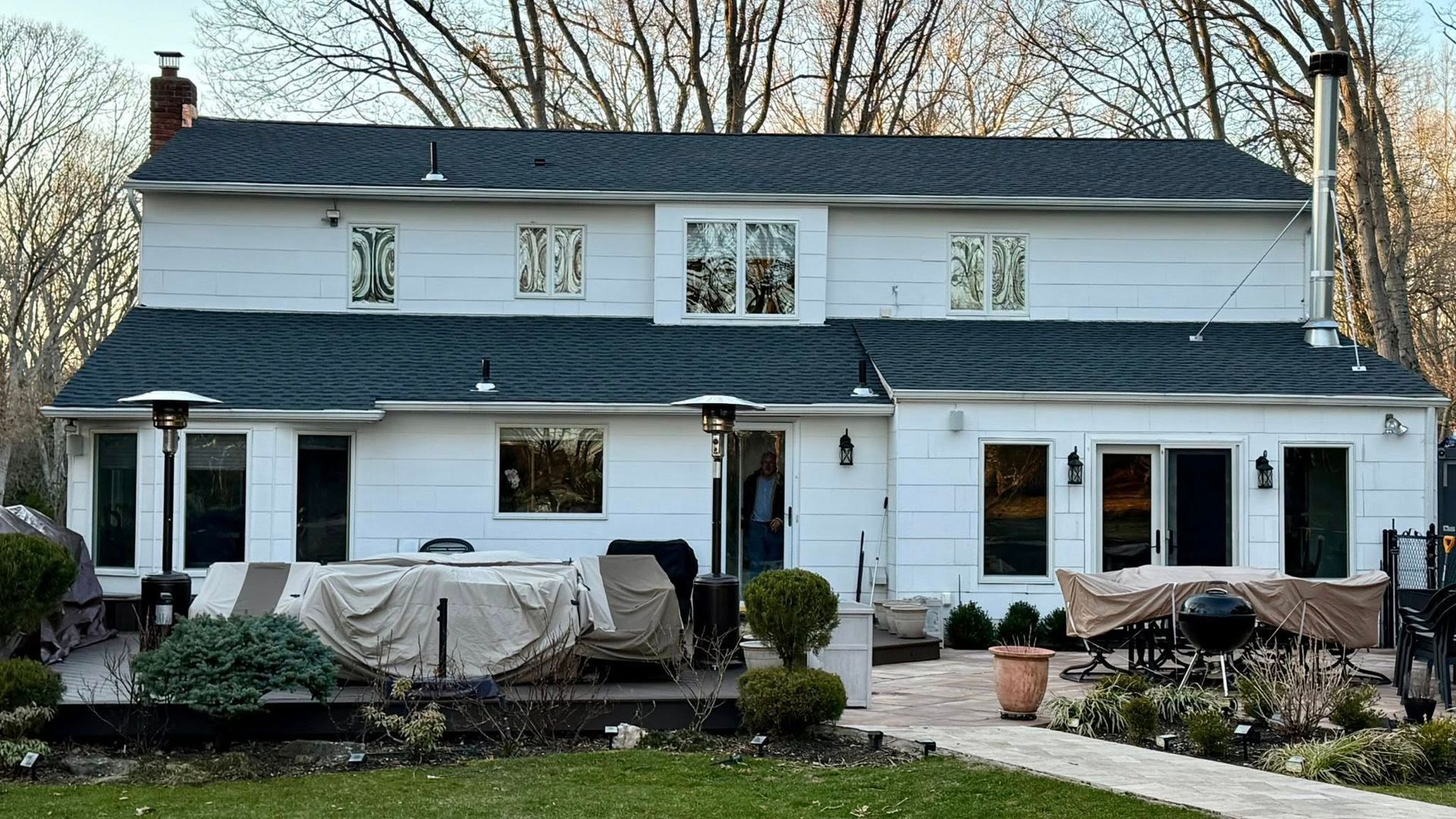 A white, two-story house with a grey roof overlooks a backyard patio area containing covered furniture and outdoor heaters.