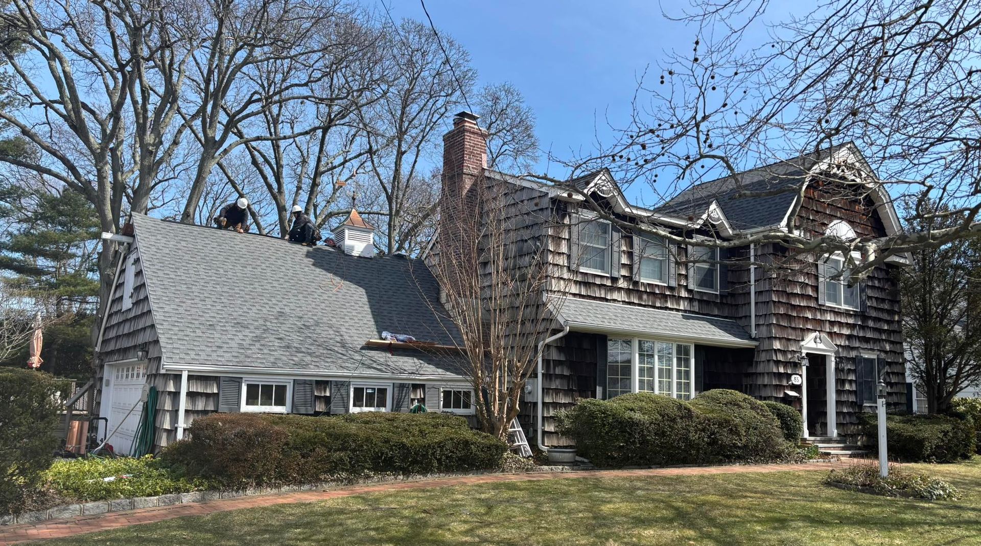 Workers on the roof of a two-story home with brown stone-textured siding and a gray shingled roof on a sunny day.