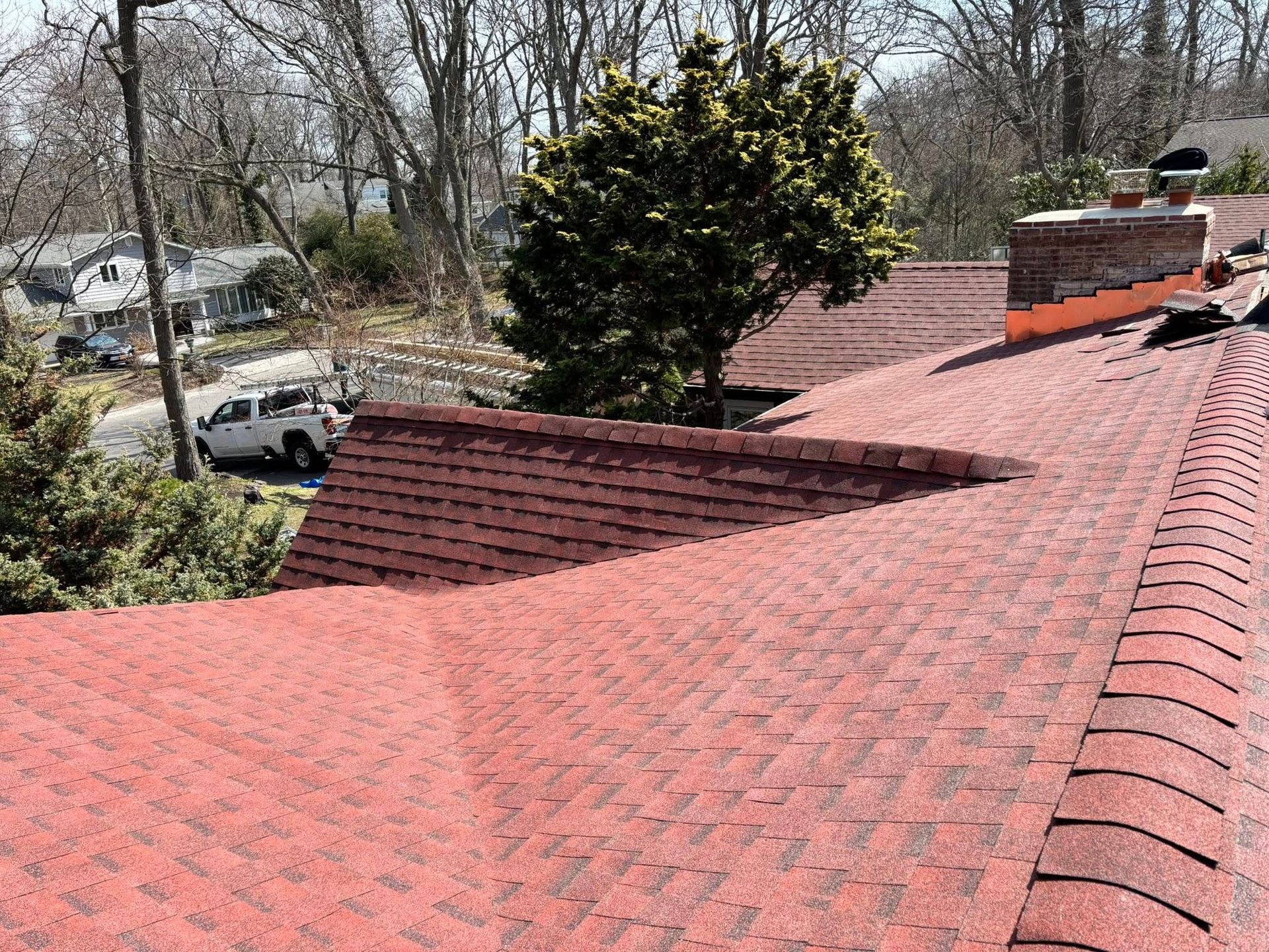 A view of red shingled roof slopes with a brick chimney and tree, overlooking a street with a parked white pickup truck.