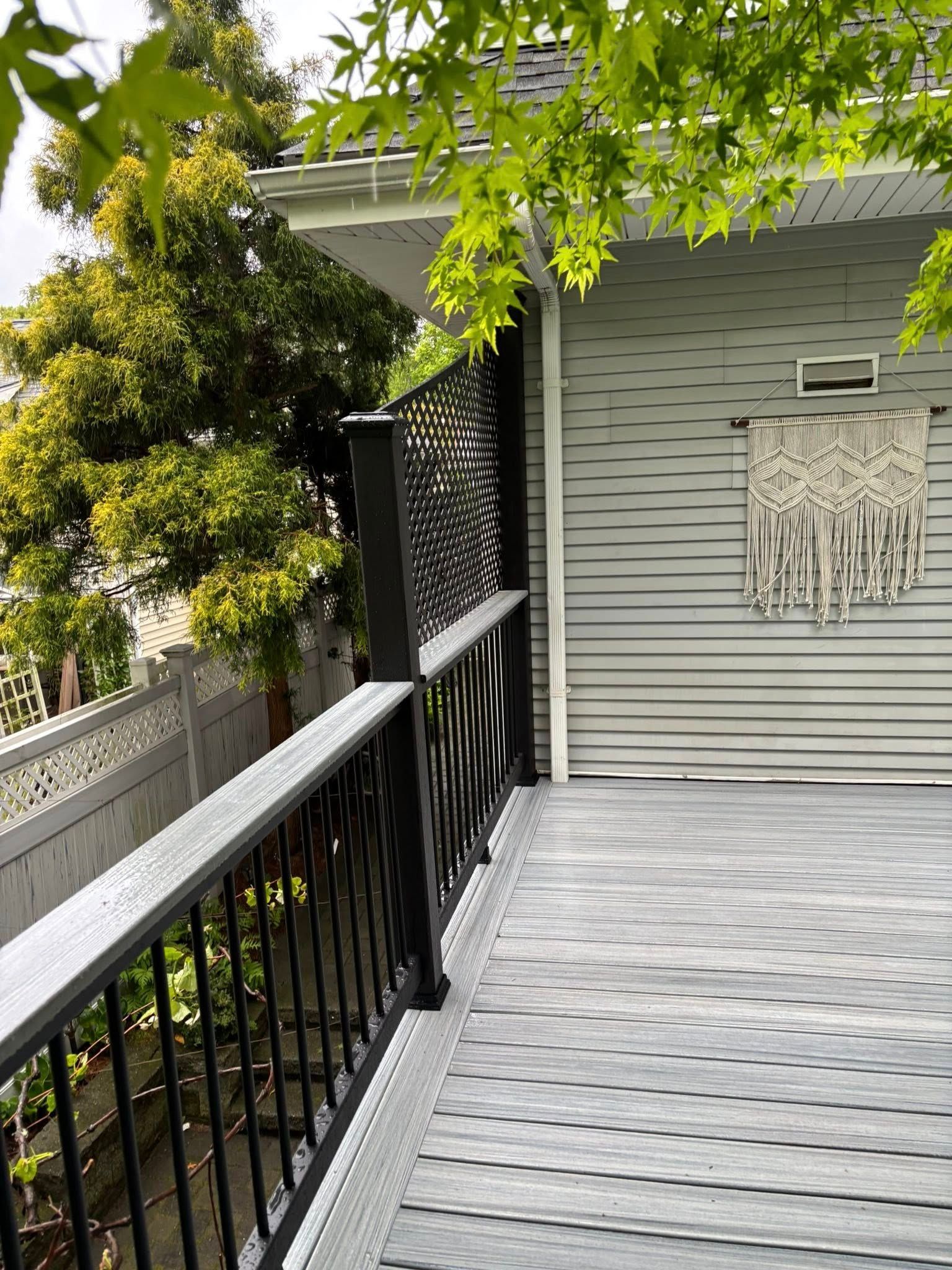 Covered porch with gray siding, black railing, and leafy trees beyond the railing
