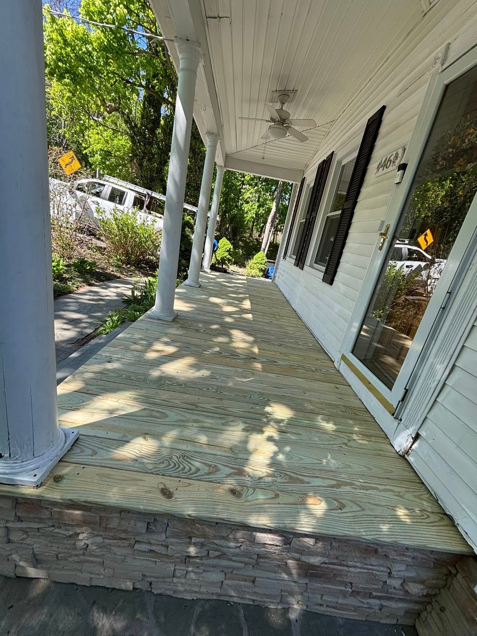 Front porch with white columns, weathered floor, and a brick step beside a house and garden path.