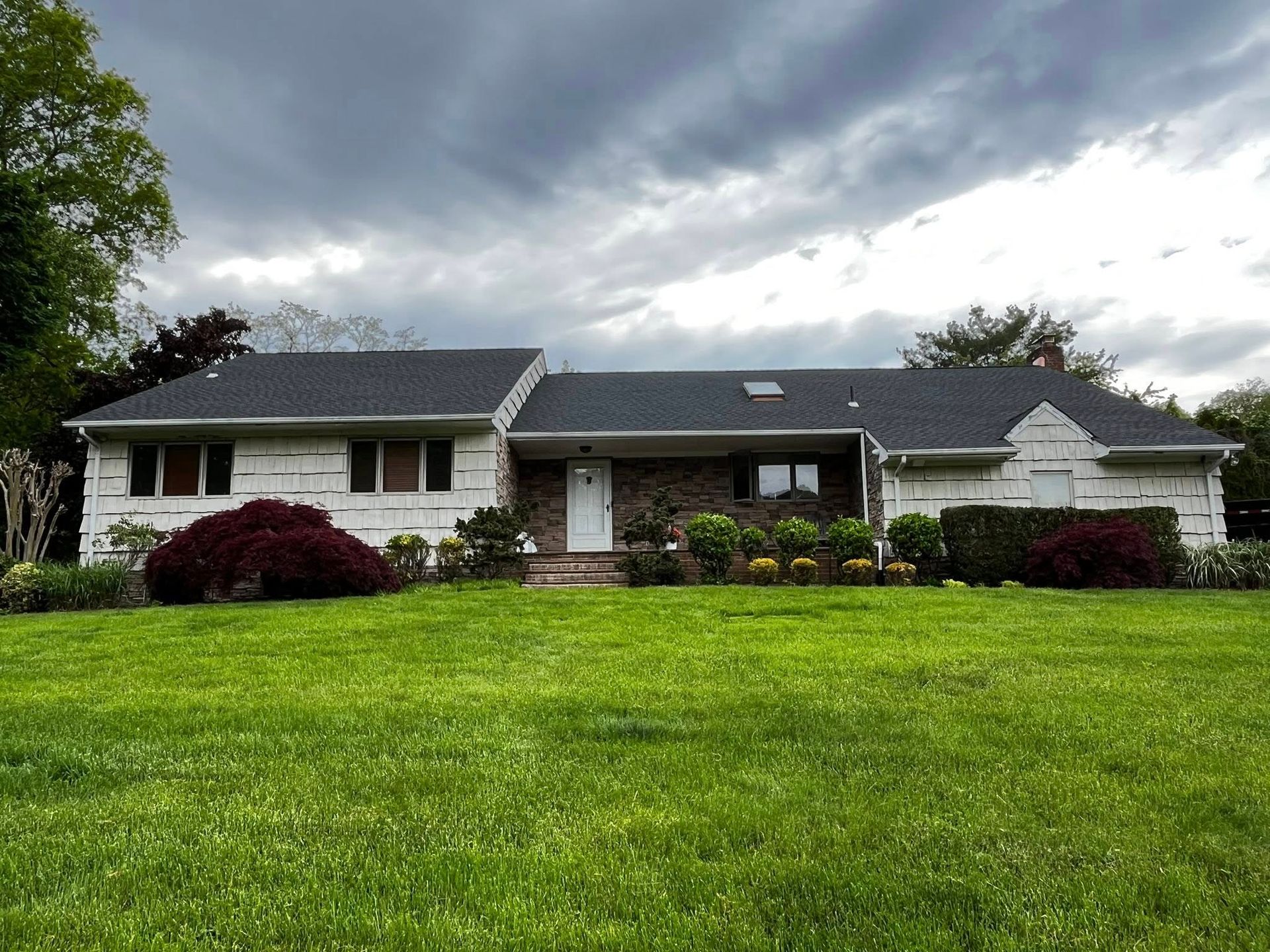 Single-story suburban house with white siding, dark roof, and a large green lawn under cloudy skies
