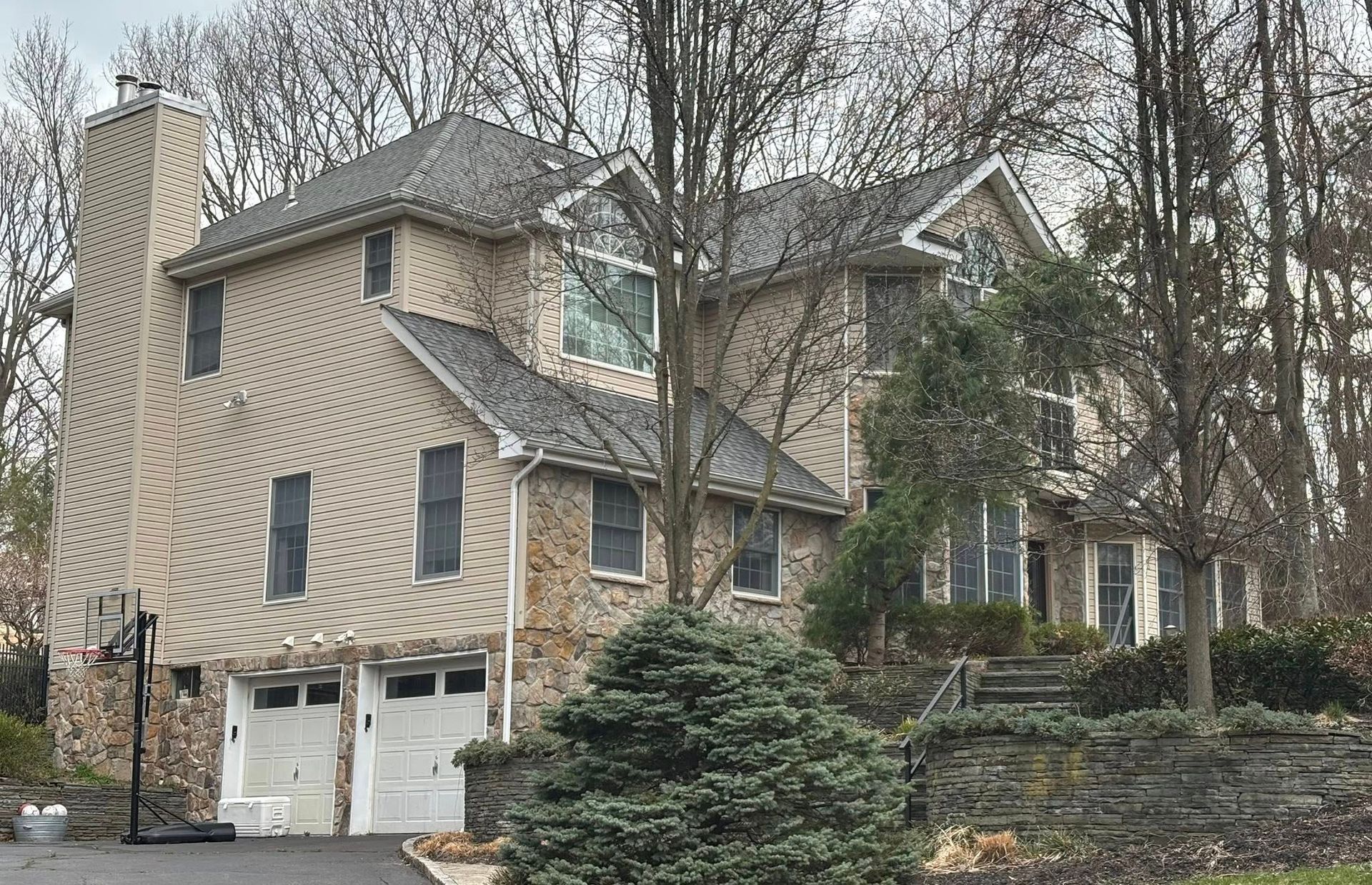 Tan house with white garage doors on a wooded hillside in winter