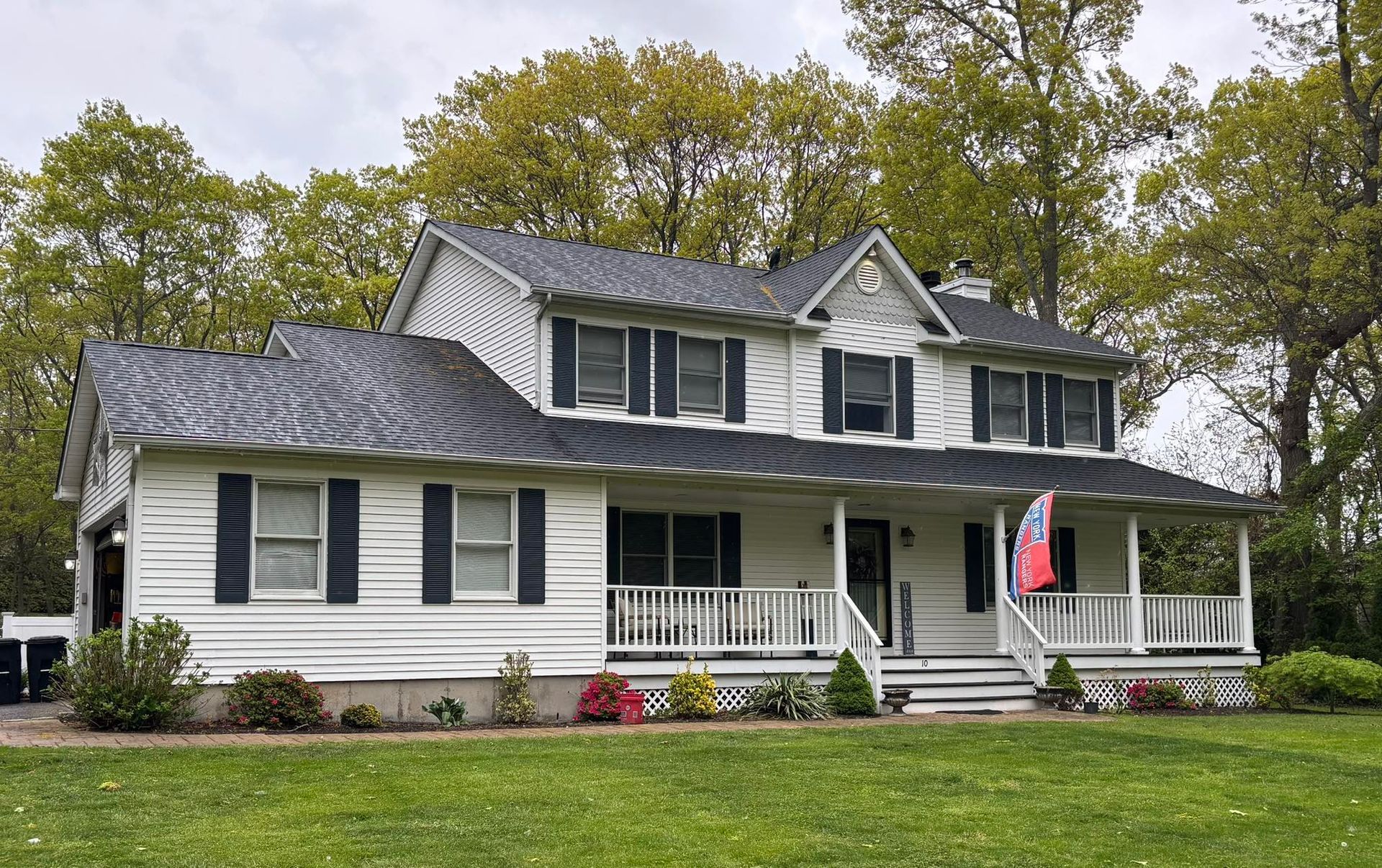 White house with dark gray roof, front porch, and American flag, surrounded by trees and flowers.