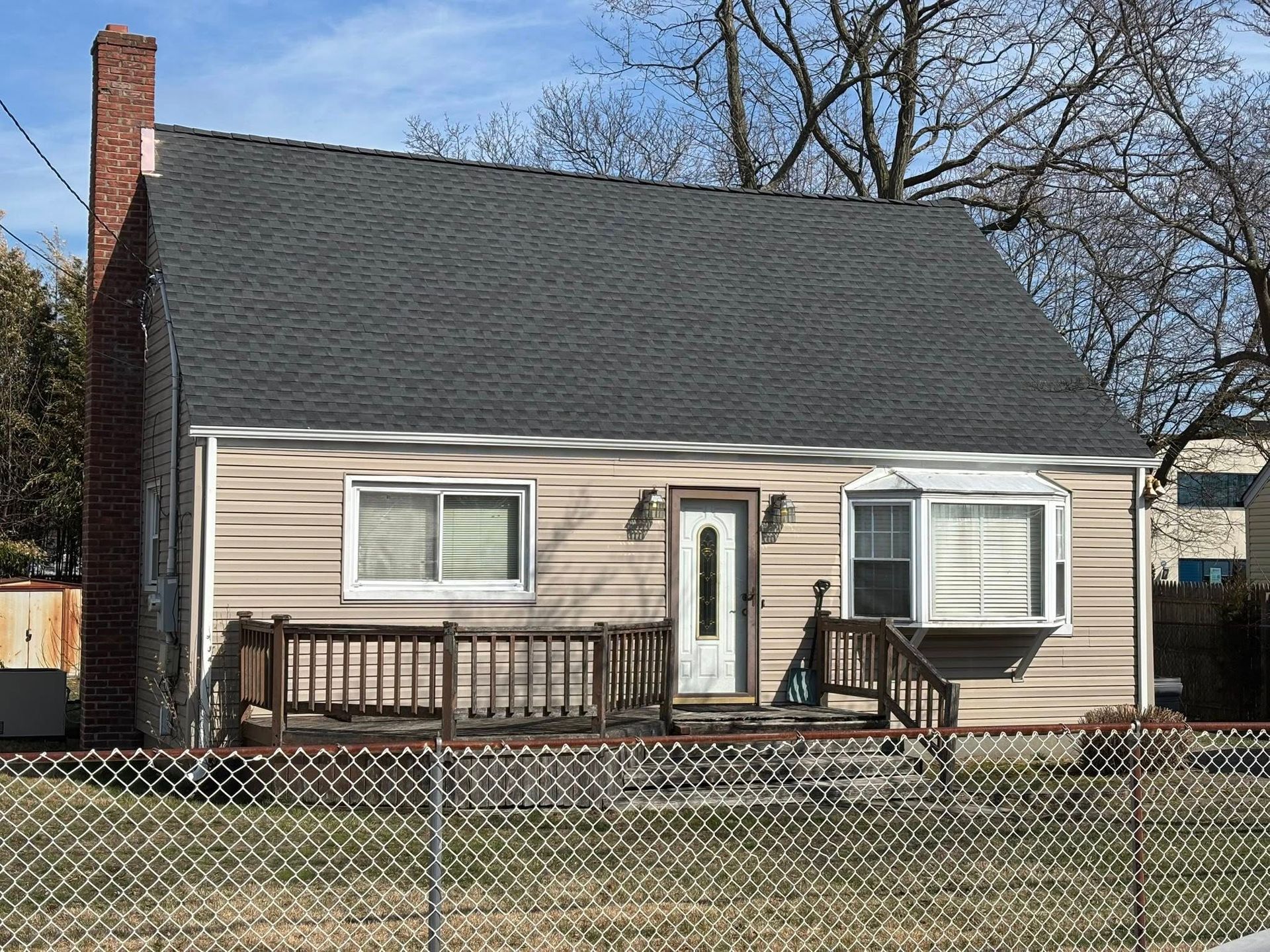 Small beige house with dark roof, front porch, and chain-link fence in a yard.