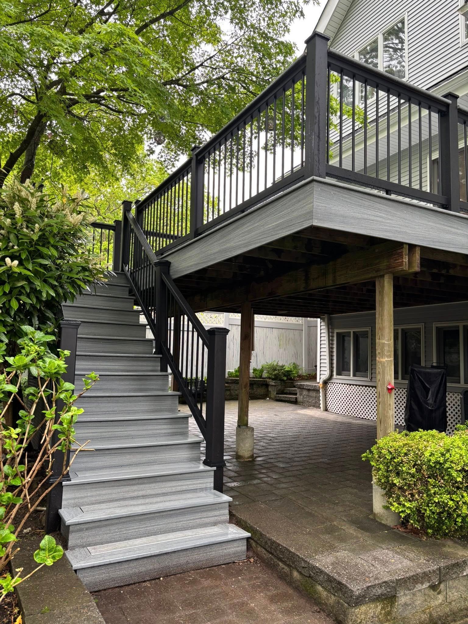 Elevated deck with gray stairs and black railing beside a house, under leafy trees