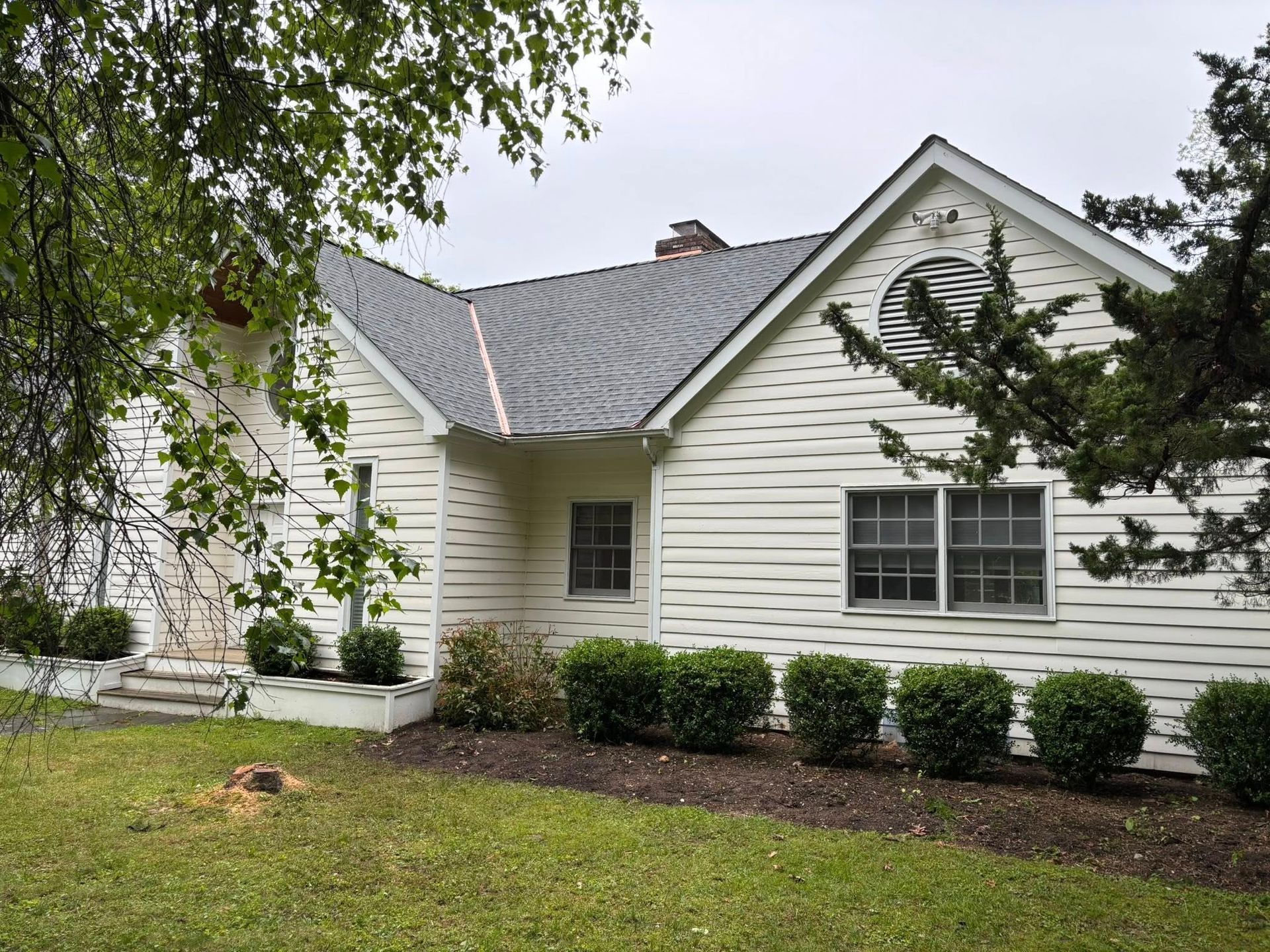 White house with gray roof and shrubs, viewed from a grassy yard under trees