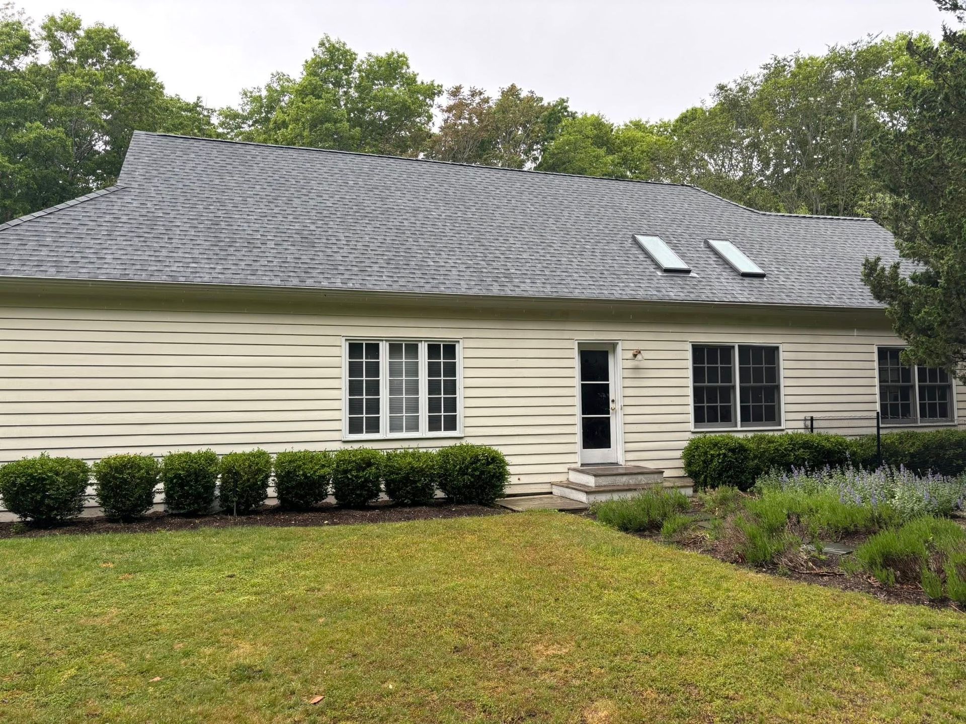 Single-story beige house with dark roof, white windows, and a front lawn surrounded by trees.
