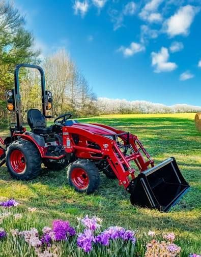Red tractor with front loader in a grassy field under a blue sky, with purple flowers in the foreground.
