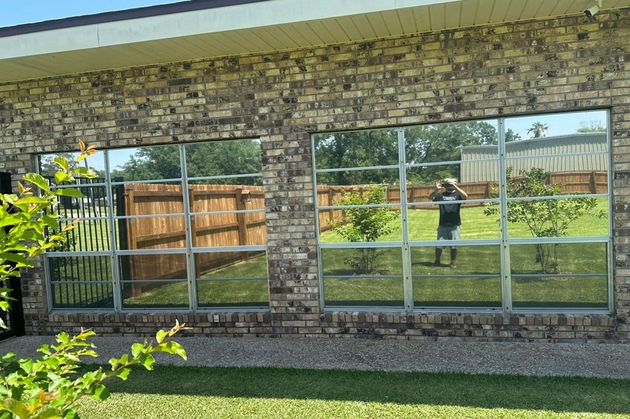 A brick exterior wall features two large windows reflecting a grassy backyard, a wooden fence, and a person taking a photo.