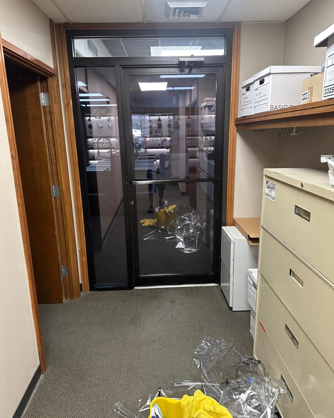 A narrow carpeted office hallway with a glass door, a metal filing cabinet on the right, and a yellow item on the floor.