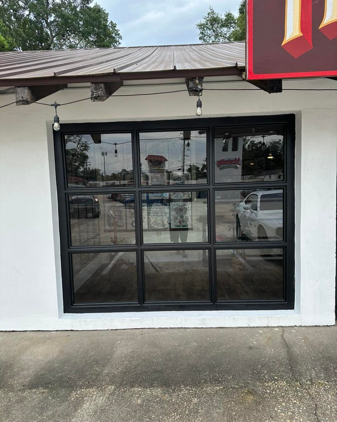 A large window with a black grid frame set into a white wall under an eave with string lights.