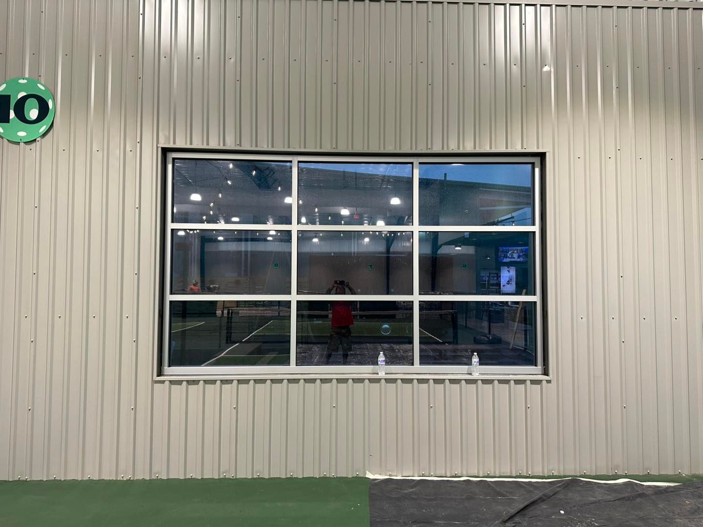 A rectangular, nine-pane window on a corrugated tan metal wall, reflecting an indoor gym area with artificial turf.