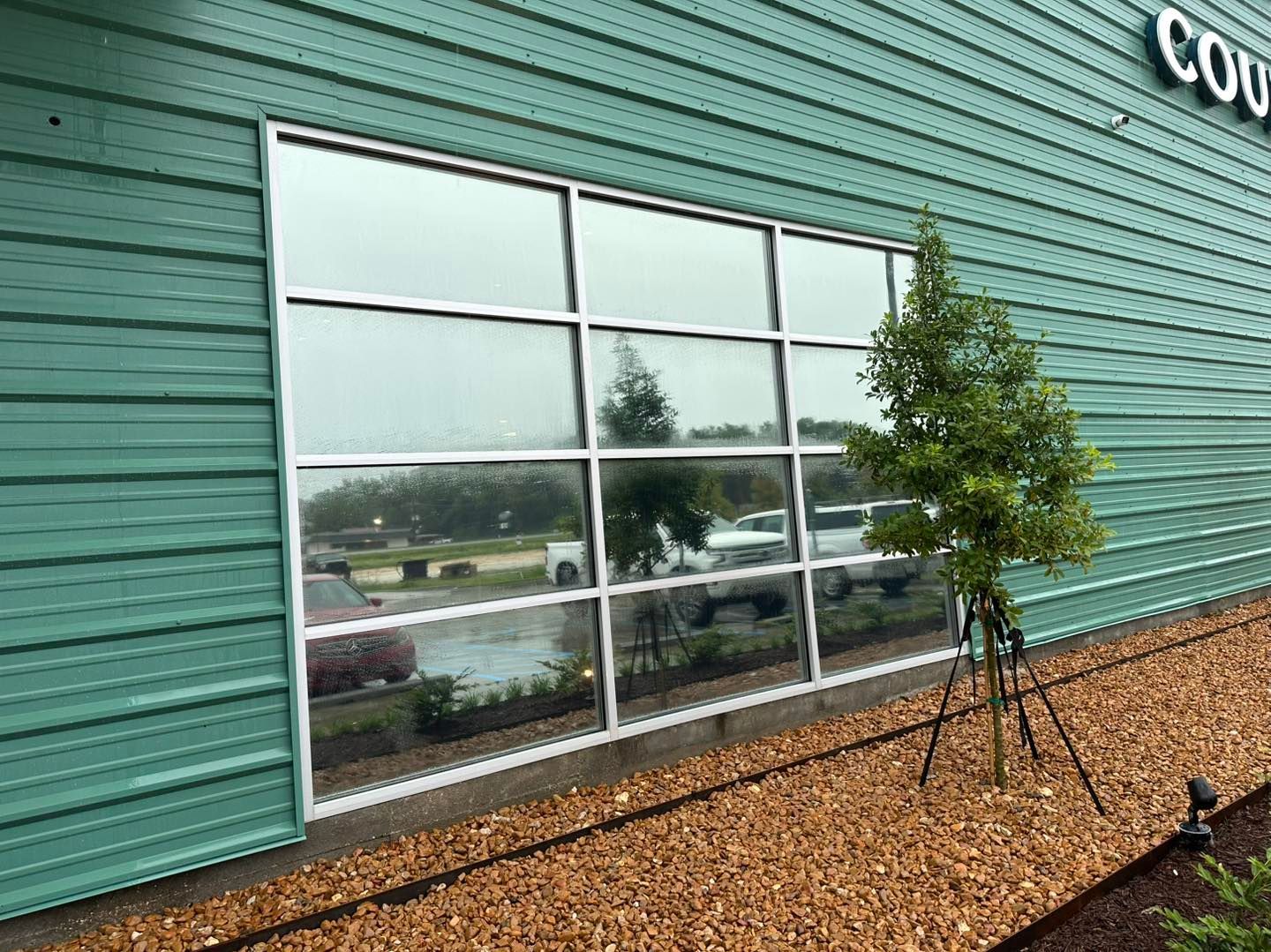 A large grid window on a teal-sided building reflects a parking lot and a small tree in a gravel landscape.