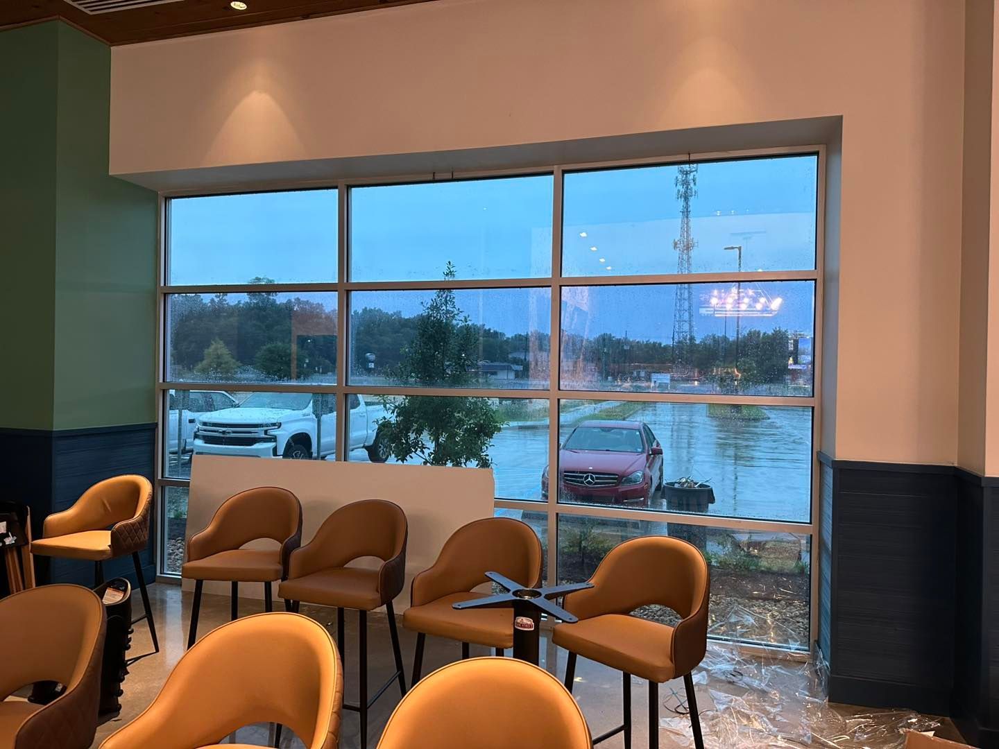 A row of tan, rounded-back chairs sits indoors against a large window overlooking a parking lot on a cloudy day.